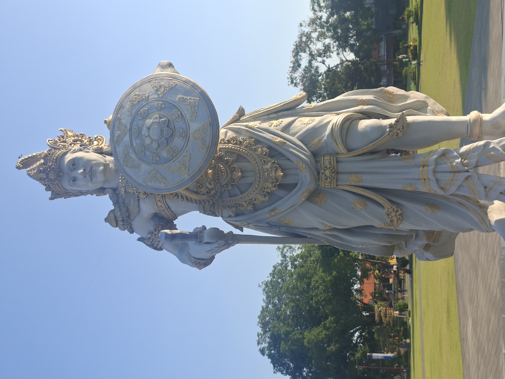 This is a waist-up, low-angle, full-color photograph of a large, grey, stone statue of a warrior or deity. The statue is adorned with intricate gold detailing on its crown, chest, and clothing. It holds a shield in its right hand and a staff or spear in its left. The statue is positioned outdoors on a paved surface, with a lush green lawn and trees in the background. The sky is a clear, bright blue. The statue is located in a park-like setting, possibly a public square or temple ground, in Gianyar, Indonesia. In the background, several small figures of people can be seen walking on the grass.