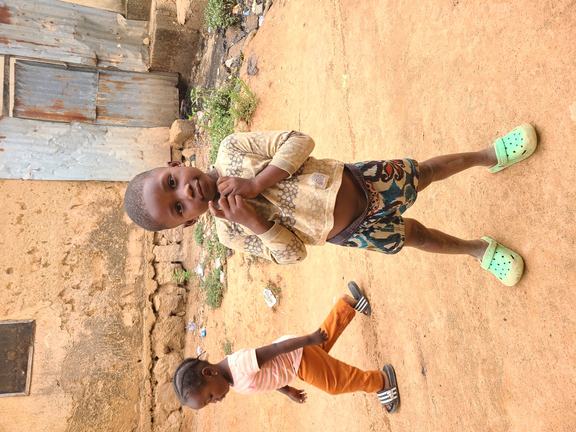 Two children stand on a dirt surface next to a building made of mud brick and corrugated metal in Jos, Nigeria. One child wears a beige long-sleeved shirt, patterned shorts, and green Crocs. The other child wears a striped peach shirt, orange pants, and black-and-white Adidas sandals. Vegetation and trash are visible near the building.