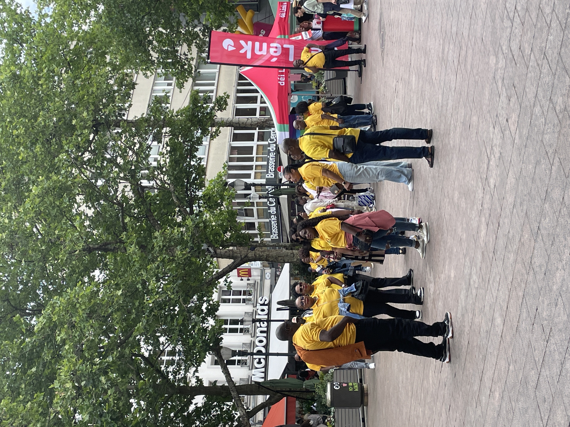 The image captures a vibrant daytime scene in the Place d'Armes, a public square in Luxembourg City, Luxembourg. Approximately 25-30 individuals, primarily adults, are visible, many of whom are wearing matching bright yellow polo shirts, indicating they are part of an organized group or event. Most people have dark complexions. They are standing in various small groups, seemingly engaged in conversations or observing the environment. One man in the foreground has an orange jacket slung over his shoulder, and another woman has a pink jacket tied around her waist. On the far right, a man in a yellow shirt holds a mobile phone to his ear.
The square's ground is paved with grey rectangular stone tiles. In the background, prominent buildings house commercial establishments. To the left, a "McDonald's" restaurant is clearly visible with its white signage and golden arches logo. Adjacent to it, a building displays the sign "Brasserie du Cercle." Further to the right, two red event tents are set up, along with a tall, vertical red banner featuring the word "Lénk" in white text and an arrow logo. Large, mature trees with dense green foliage are dispersed across the square, their branches extending across the upper portion of the image, partially obscuring the upper floors of the background buildings, which are light-colored with multiple windows. A dark grey cylindrical public waste bin with a graphic of a smiling face is visible on the left side of the frame. The overall lighting suggests an outdoor daytime setting, possibly under an overcast sky.