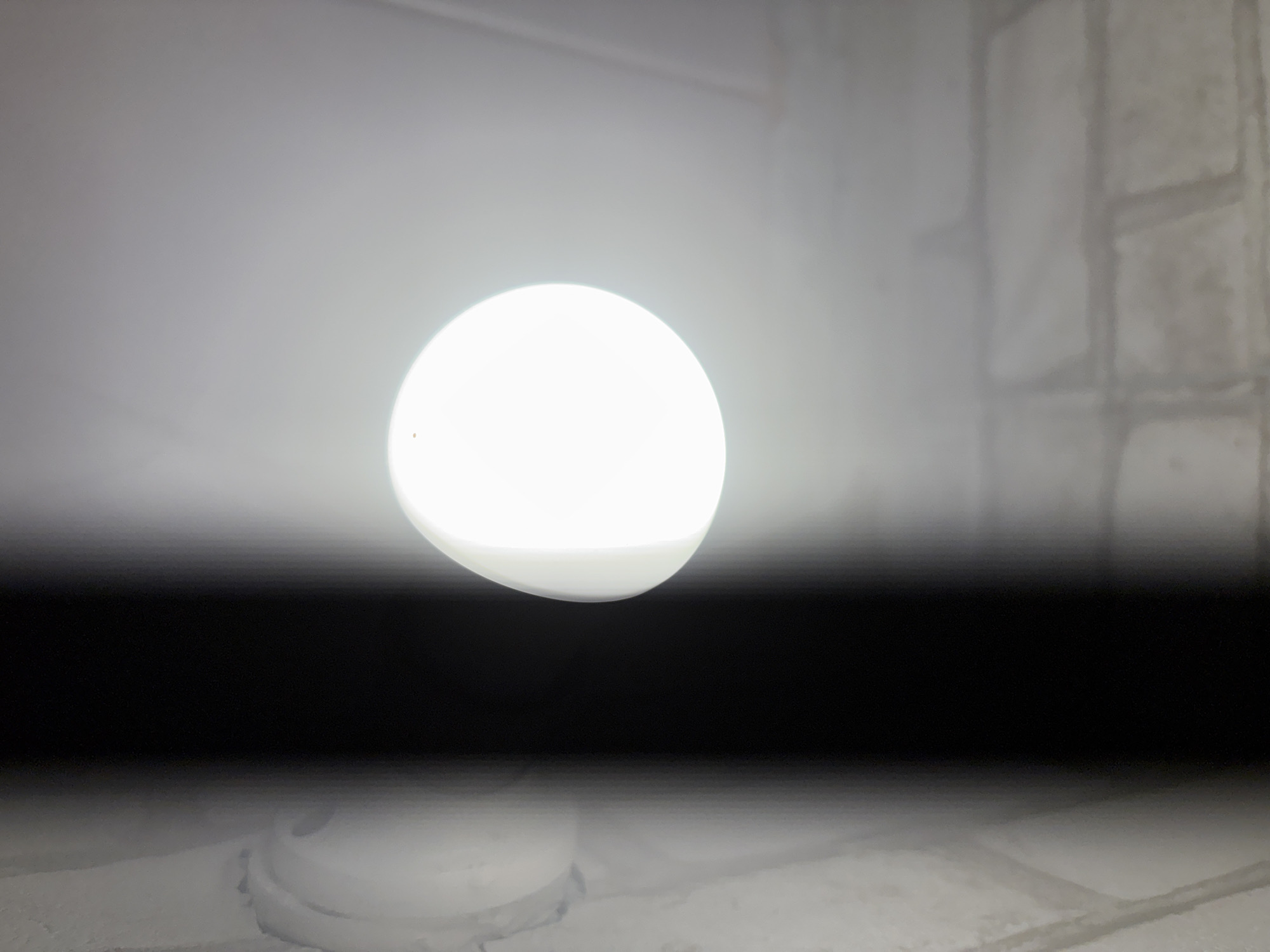 An overhead shot shows a bright, round, white LED light fixture emitting a strong glare. The light is positioned on a ceiling, which appears to be textured and possibly painted white. To the left of the light, a dark, vertical shadow or beam cuts across the frame. The background consists of a brick wall with a light-colored, textured finish. The overall scene suggests an indoor environment, possibly a room or hallway, with a strong light source.