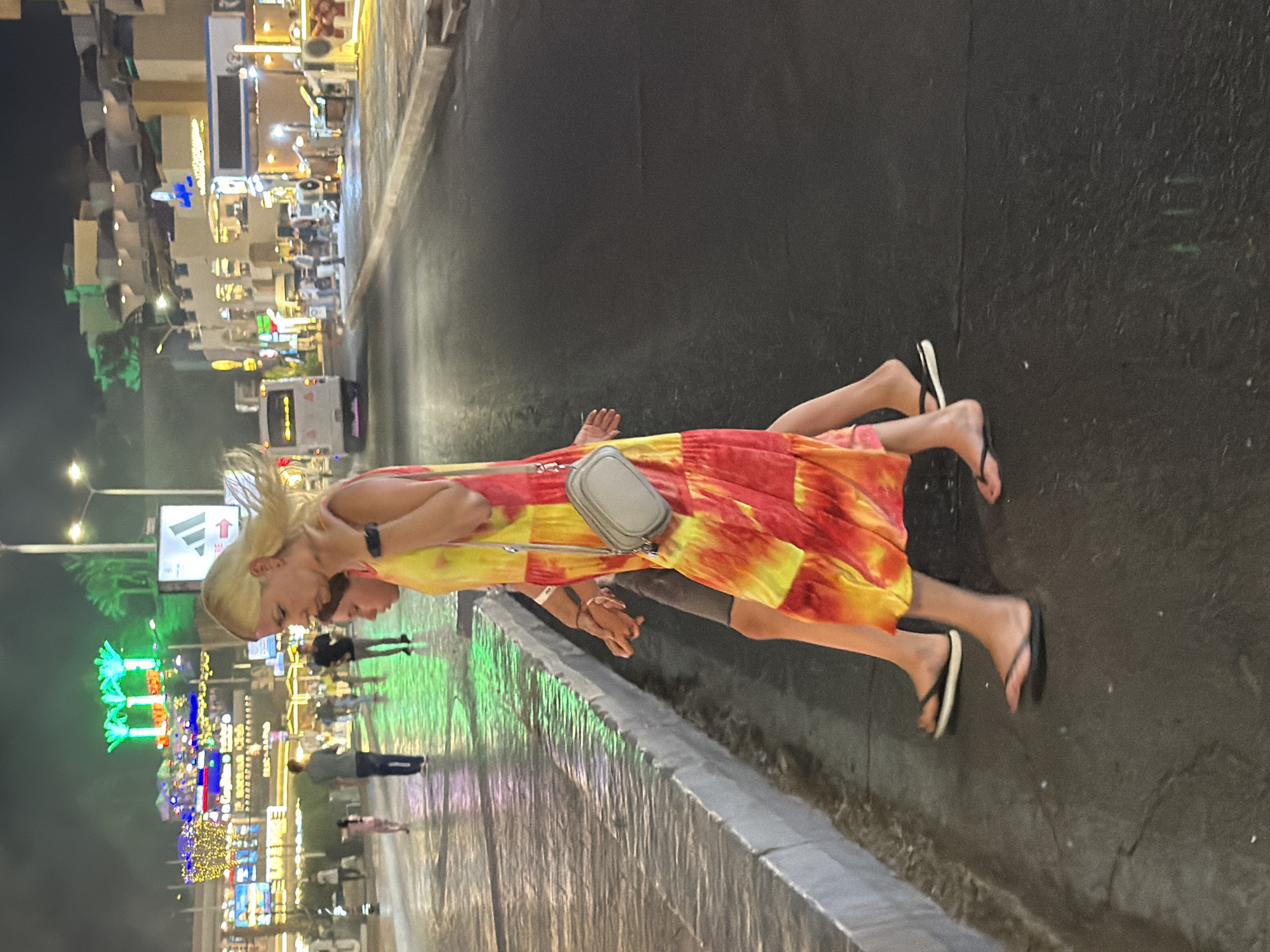 A medium shot captures a woman and a child walking side-by-side at night on a wet, dark asphalt street in Sharm El Sheikh, Egypt. The woman, in the foreground on the right, wears a vibrant tie-dye dress in shades of yellow, red, and orange, and a small light-gray crossbody bag. She is wearing flip-flops. The child, to her left and slightly behind, is wearing a dark grey t-shirt and shorts, also in flip-flops. Their hands are clasped. The street is illuminated by neon signs and lights from buildings on the left, which are decorated with festive lights, and further down the street on the right. Palm trees with green illumination are visible in the background on the left. A white bus is parked on the right side of the street. The lighting creates reflections on the wet pavement, suggesting recent rain or a fountain.
