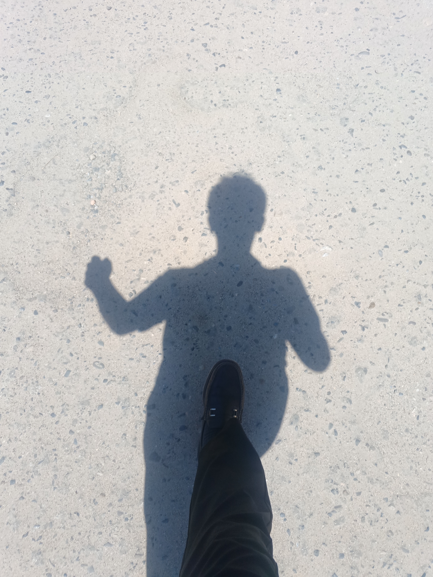 The image captures a shadow of a person walking on a concrete surface, likely in the city of Kusaho, Uzbekistan. The shadow is projected onto a light-colored, coarse concrete ground, speckled with small dark and light aggregates. The person's shadow shows a figure with a raised right arm, fist clenched, as if in a pose of strength or determination. The lower portion of the shadow is cast by a leg wearing dark trousers and a dark shoe. The perspective is from a high angle, looking down at the ground and the shadow. The environment appears to be an outdoor, possibly a pedestrian walkway or an open area with a concrete ground. The lighting suggests a sunny day, casting a distinct shadow.