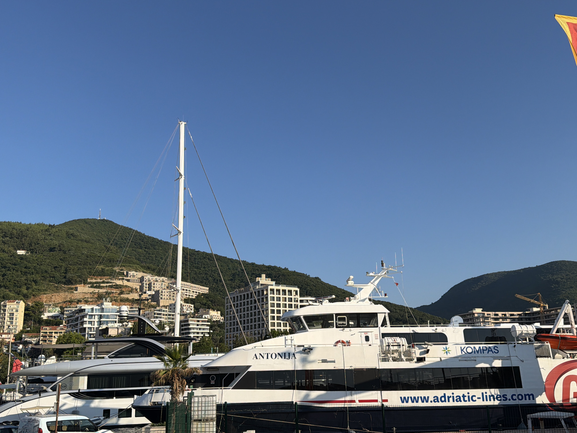 A wide shot captures the clear blue sky above the waterfront in Budva, Montenegro, under bright sunlight. In the foreground, a white ferry named "ANTONIJA" is docked, with the text "KOMPAS" and the website "www.adriatic-lines.com" visible on its side. Behind the ferry, other boats and yachts are moored. The backdrop features lush green hills dotted with buildings, indicating an urban setting on the coast. A tall mast from a sailboat is prominent on the left, and a red and yellow flag is visible in the upper right corner. The scene suggests a typical day at a busy port.