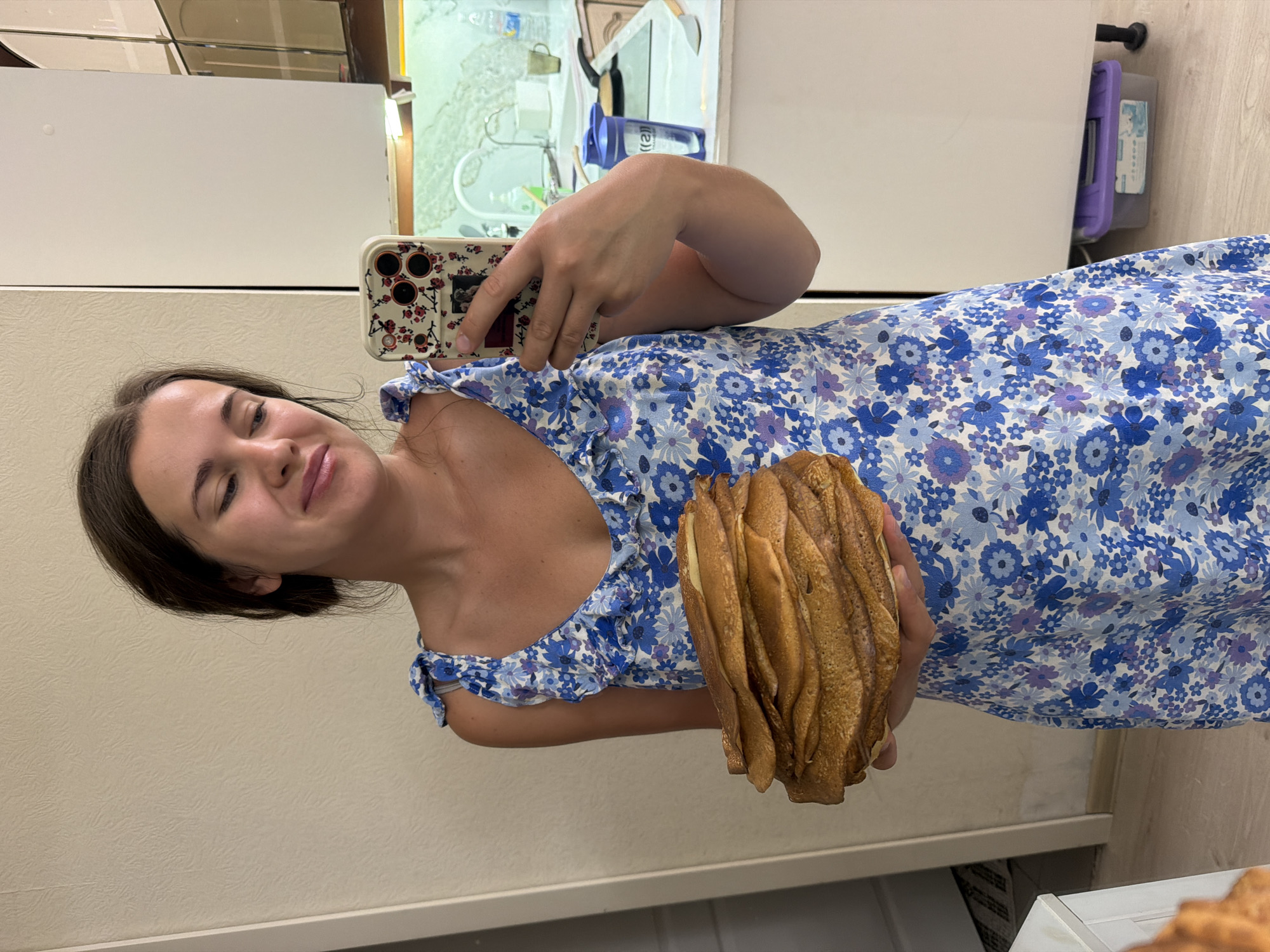 A young woman stands in a brightly lit kitchen, holding a large stack of crepes. She is wearing a blue floral dress and has her hair pulled back. She is taking a selfie with her phone, which has a floral case. The kitchen appears to be in a residential apartment in Sochi, Russia, with white cabinets, a granite countertop, and a tiled backsplash. There is a blue water bottle and a pan on the stove in the background.