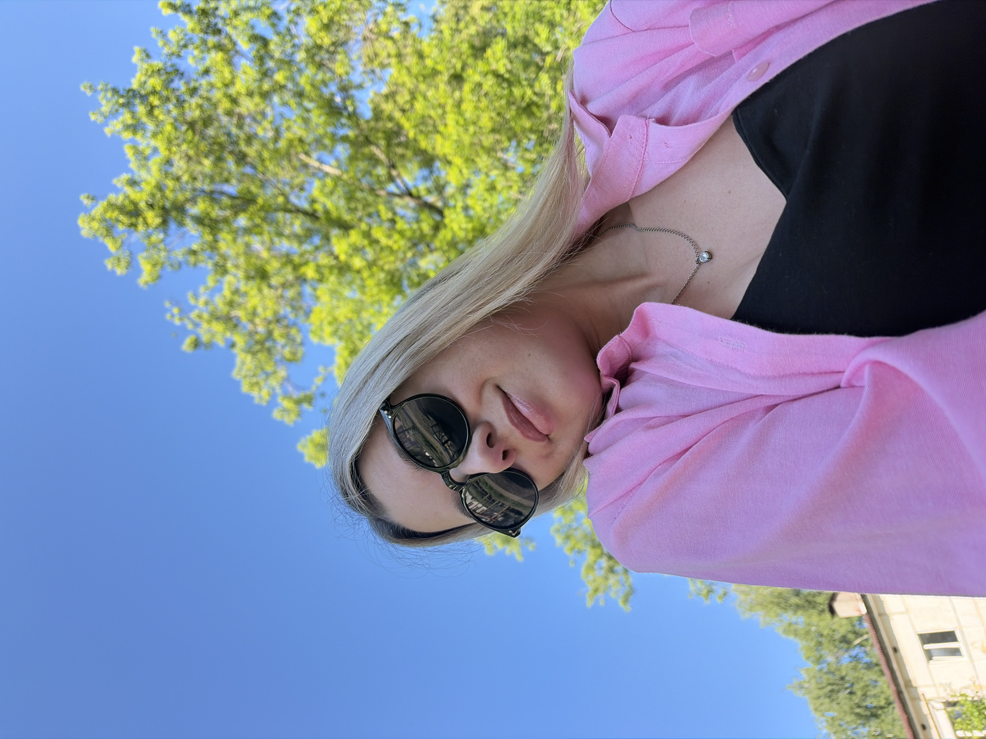 A young woman with blonde hair and sunglasses is looking up towards the camera. She is wearing a pink button-down shirt over a black tank top and a silver chain with a heart-shaped pendant. The background is a clear blue sky with a large, leafy green tree. To the left, a building with a light-colored facade and a chimney is visible. This scene is likely in the city of Chaikovsky, Russia, judging by the surrounding environment which includes typical Russian urban architecture and vegetation.