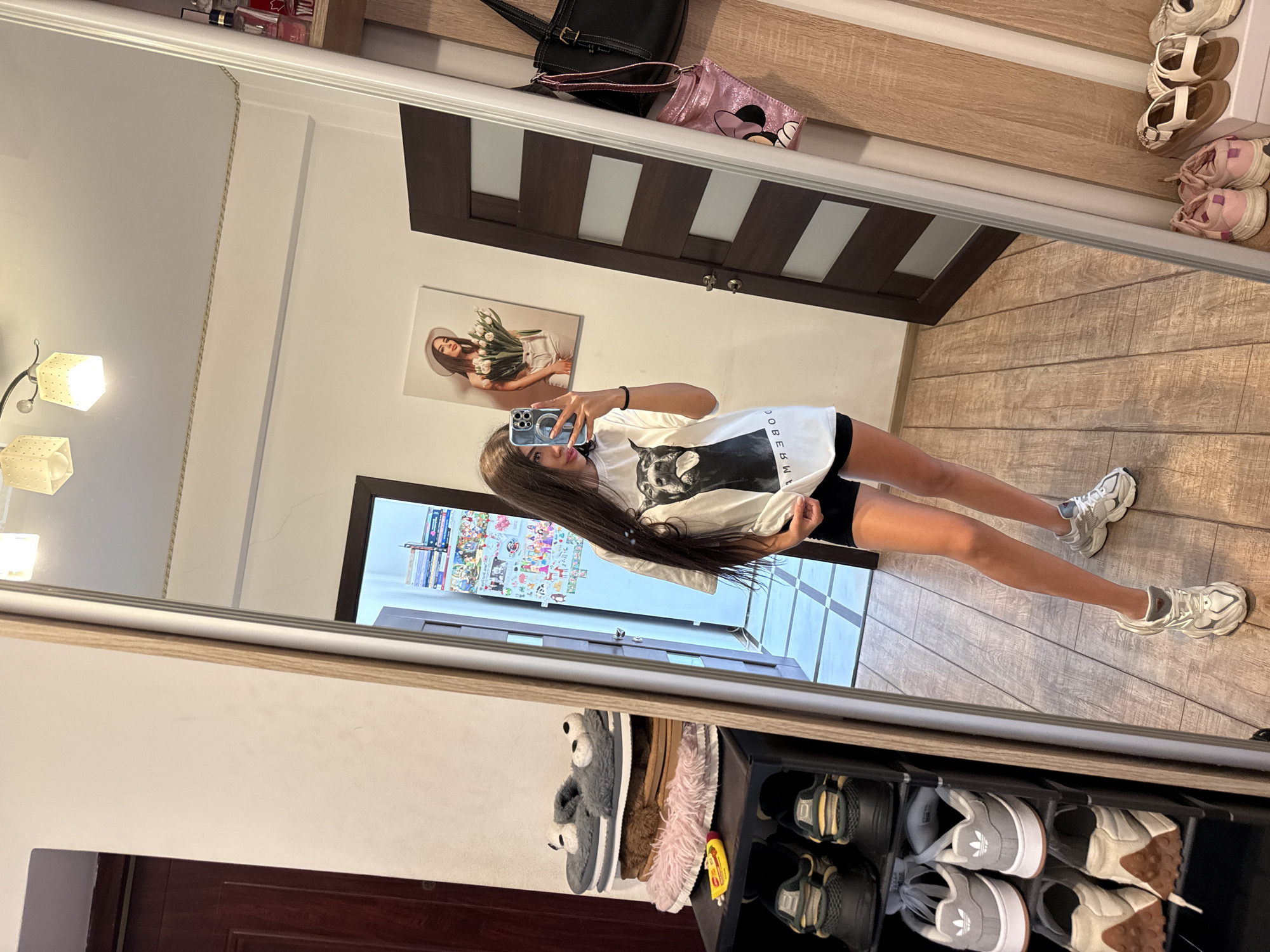 The image depicts a young woman with long, dark hair taking a mirror selfie in what appears to be a residential hallway or bedroom in Chișinău, Moldova. She is wearing a white t-shirt with a black and white graphic of a dog and black shorts. Her reflection shows her holding a smartphone and a black bracelet on her wrist.
The environment includes a large mirror dominating the frame, reflecting the woman and the hallway behind her. To the left of the mirror, a narrow, dark brown wooden door with glass panels is visible. To the right of the mirror, a wooden closet or shelving unit is partially seen, with various items including handbags and shoes displayed. On the wall to the left of the door, a framed picture of a woman holding tulips is hanging. A shoe rack filled with sneakers and slippers is positioned in the foreground to the left, and additional shoes are visible on the floor in the lower right corner. The floor is made of light brown wooden planks. The lighting suggests an indoor setting with overhead lighting fixtures visible in the reflection.