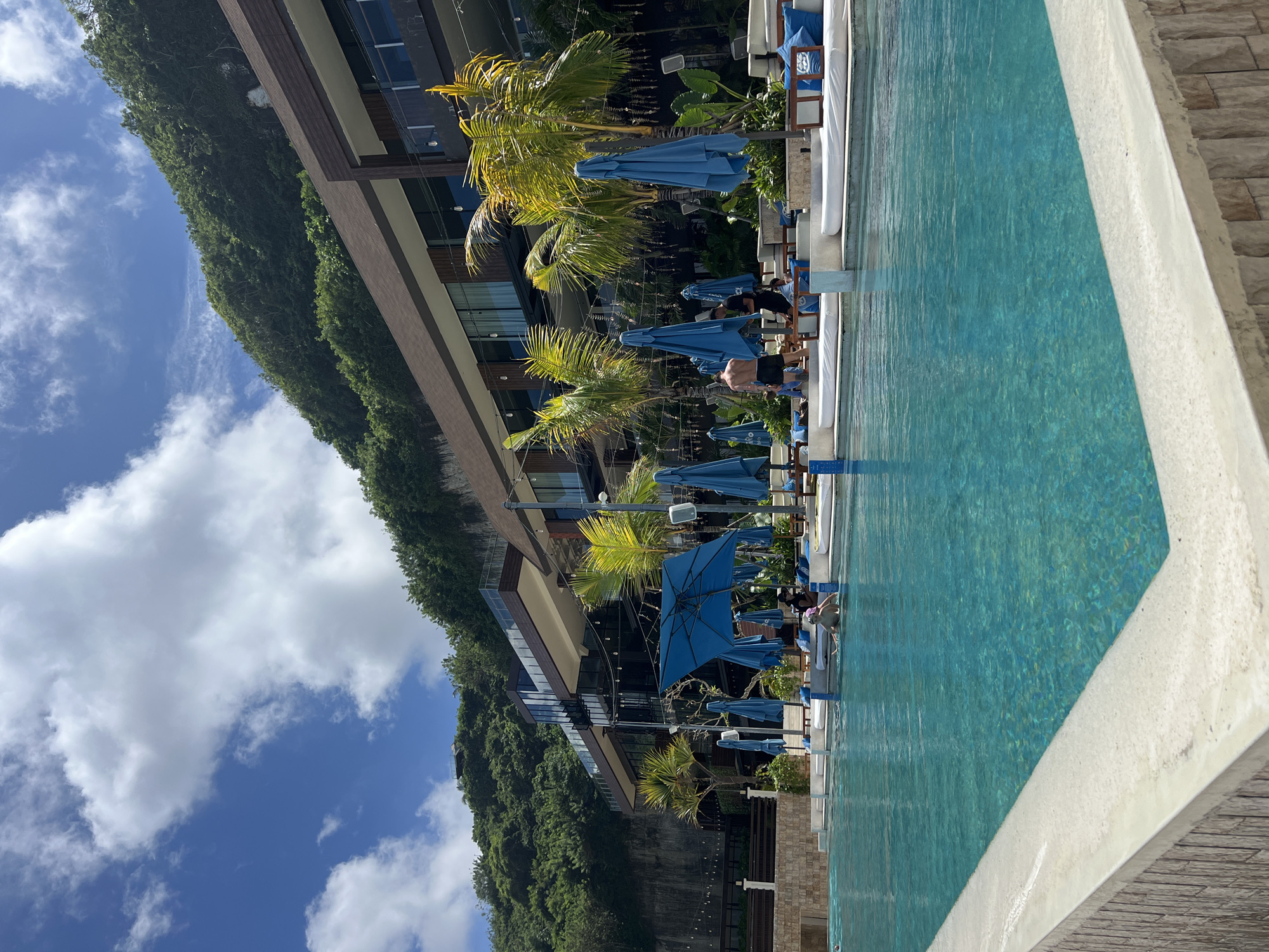 A vibrant outdoor scene captures a resort pool area under a bright, sunny sky in Kuta Selatan, Indonesia. In the foreground, a large, inviting turquoise-blue swimming pool dominates the right side of the frame, bordered by a light-colored concrete edge. Several people are enjoying the amenities; one individual is partially submerged in the pool, while others are seated on white lounge chairs under numerous bright blue sun umbrellas lining the pool deck.
To the left of the pool, a modern, multi-story building with dark-framed windows and balconies stands, likely part of the resort. Behind this structure, a steep, lush green hillside covered in dense tropical foliage extends into the distance, suggesting a scenic, natural setting. Palm trees are scattered around the pool area, enhancing the tropical feel. The sky above is a clear, brilliant blue adorned with scattered white cumulus clouds. The scene conveys a relaxed, leisurely vacation atmosphere on a bright, sunny day, likely midday or early afternoon, with clear shadows indicating strong sunlight. No visible text is present.