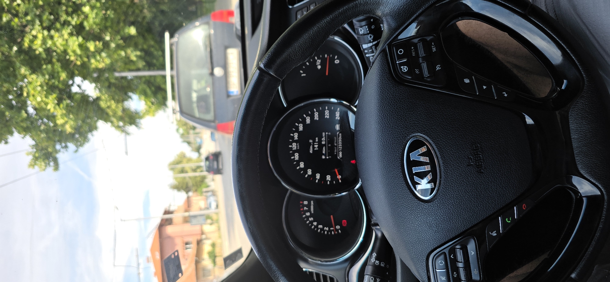 The image is a first-person perspective from inside a car, showing the dashboard and steering wheel. The steering wheel has the KIA logo in the center and various buttons on its sides. Two gauges are visible: a tachometer on the left and a speedometer on the right, which displays a speed of 141 km/h and an odometer reading of 123390 km. The car is in motion on a street with trees and buildings visible outside the windshield. Another car is in front, and the sky is cloudy.