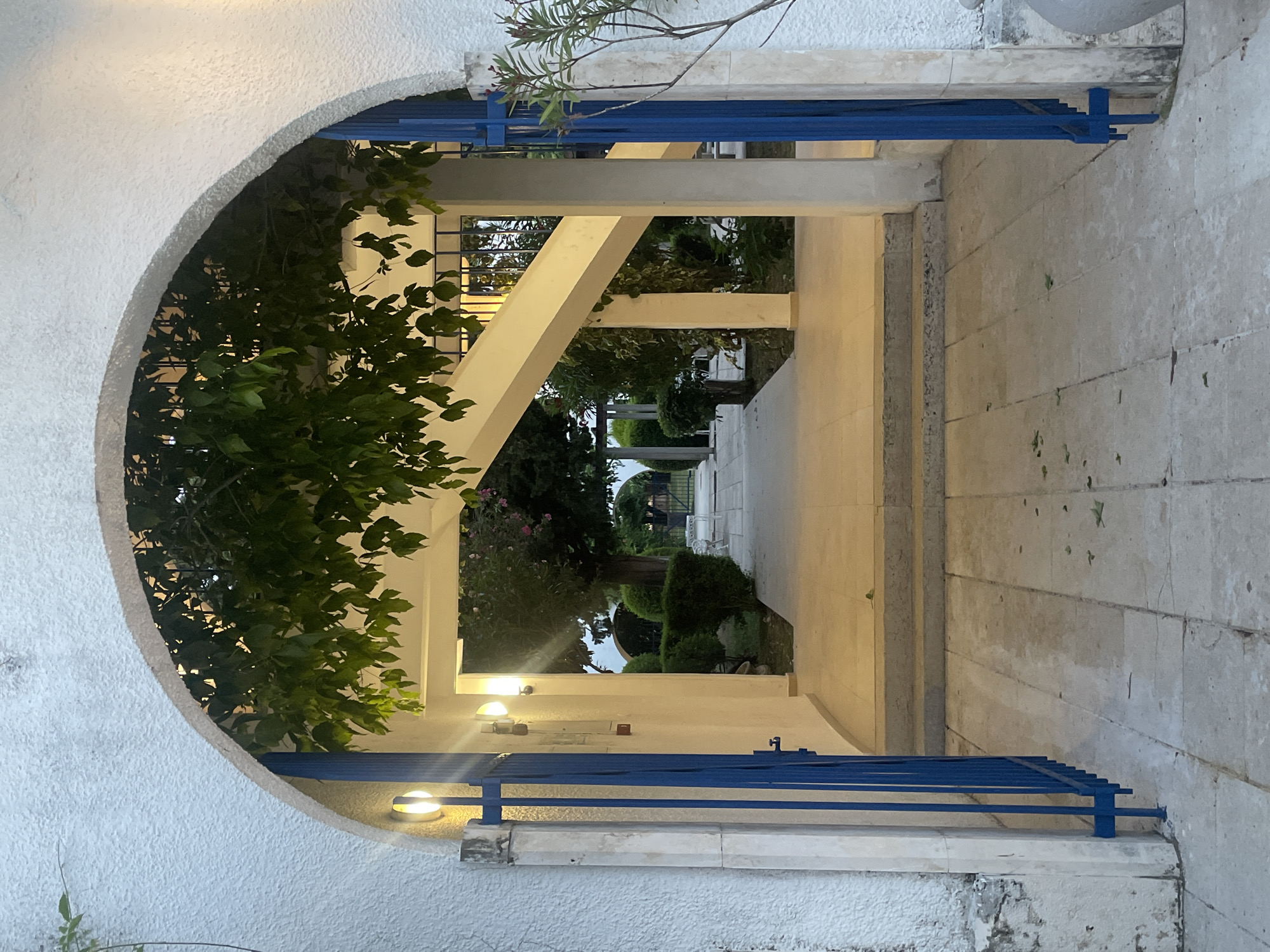 The image presents a tranquil view through an arched passage at what appears to be dusk or early evening, indicated by the active artificial lights. The foreground is dominated by a thick, textured white archway, which frames the scene beyond. Within this arch, on both the left and right sides, are vibrant blue metal railings or gates, adding a distinct color accent.
To the immediate left of the archway, dark green foliage from a plant or tree spills into the opening, creating a natural, slightly overgrown feel. Beyond the arch, a paved pathway extends into the distance, guiding the eye through a structured outdoor space. This pathway is flanked by light-colored architectural elements, including rectangular pillars and beams that support an overhead structure, creating a sense of depth and perspective.
Several wall-mounted lights, emitting a warm yellow glow, are visible along the left side of this inner passage, illuminating the walkway. The passage eventually opens into a garden area, where dark green trees and manicured bushes are visible under the fading light. Hints of an outdoor seating area can be discerned further in the background. On the far right of the scene, a textured concrete or stone wall rises alongside the passage. The overall impression is that of a peaceful and inviting outdoor environment, possibly within a resort, courtyard, or large garden. There are no people or visible text in the image.