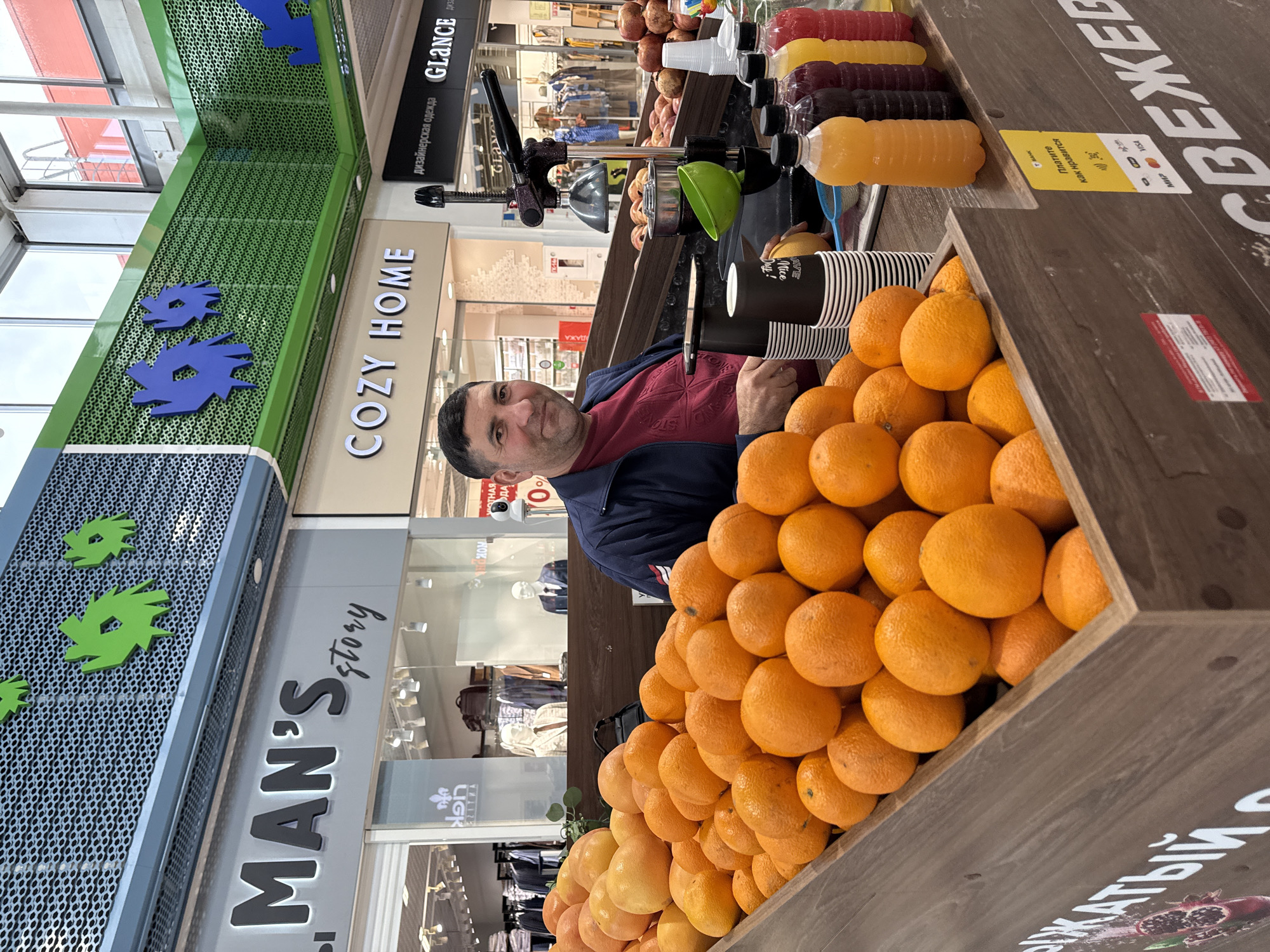 The image depicts a man standing behind a fruit stand filled with oranges and other fruits, likely in a marketplace or shopping mall in Nizhny Novgorod, Russia. The man, who appears to be the vendor, is wearing a dark blue jacket over a maroon shirt. He is smiling and looking towards the camera. In front of him, a juice press is set up, with cups and bottles of juice lined up. The background shows storefronts with signs, including "MAN'S story" and "COZY HOME." The overall scene suggests a commercial environment where fresh juice is being sold.