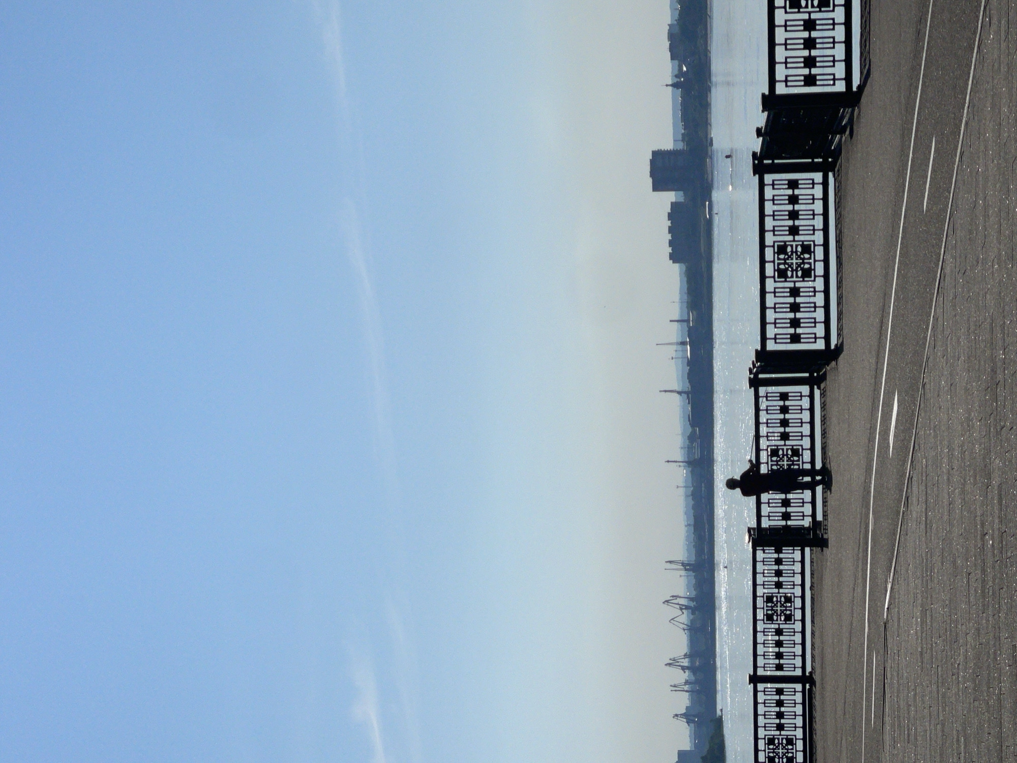 A lone figure stands at a decorative metal fence overlooking a wide, calm river on a clear, bright day. The figure, silhouetted against the water, appears to be looking out towards a distant city skyline. The skyline features industrial cranes and several buildings. The foreground shows a paved walkway with white lines, suggesting a road or bike path. The overall impression is one of quiet contemplation against an urban and industrial backdrop. The location is likely the embankment of the Yenisei River in Krasnoyarsk, Russia, specifically the area near the Krasnoyarsk Hydroelectric Power Station Dam, given the presence of industrial cranes and the characteristic wide river.