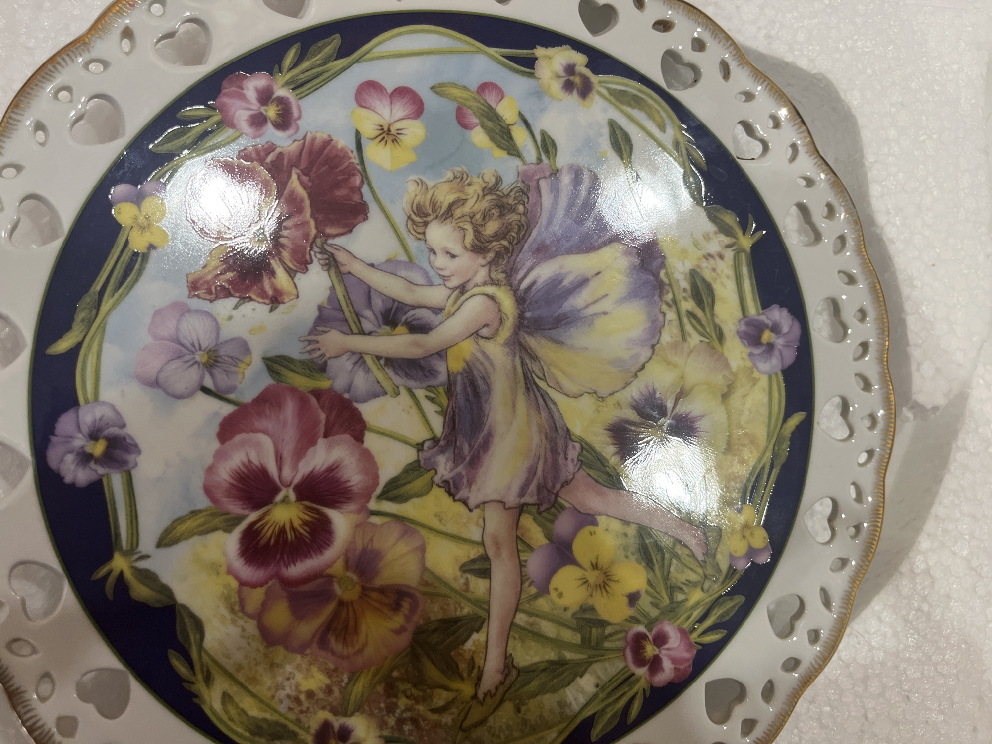 The image shows a decorative plate with a painted scene. The scene depicts a young child with blonde curly hair wearing a purple and yellow dress, appearing to be dancing or flying amongst large, colorful pansies. The child is reaching up towards a large red and yellow pansy. The background of the plate is a light blue sky. The plate has a dark blue border around the painted scene and a white, lacy edge with heart cutouts. A thin gold trim runs along the outer edge of the plate. The plate is resting on a white, textured surface. The location is unknown.