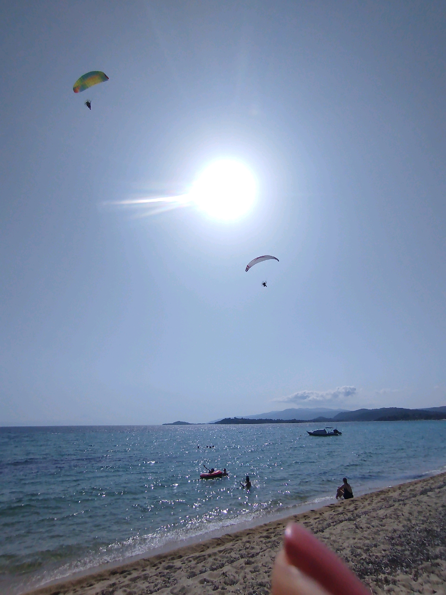 This image captures a sunny day at the beach in Sithonia Municipal Unit, Greece. The sky is bright blue with the sun prominently featured and creating a glare. Two paragliders are visible in the sky, one in the upper left with a colorful wing and the other in the mid-right with a white and red wing. The ocean is calm with visible waves breaking on the shore. Several people are in the water, some swimming and one on a red inflatable raft. A small boat with a canopy is anchored in the distance. The beach is sandy, with some people sitting on the sand near the water's edge. The foreground is partially obstructed by a blurred finger.