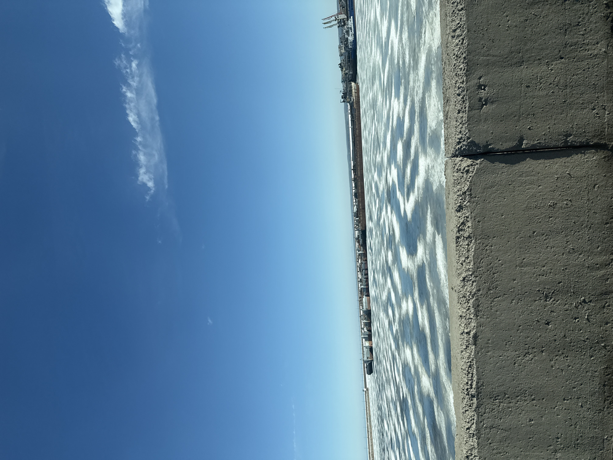 The image shows the Kama River from Chaikovsky, Russia. The picture is taken at an elevated angle, looking across the water. In the distance is an industrial port with cargo ships and cranes. The foreground reveals the corner of a gray, textured concrete structure, possibly a pier or embankment. The river surface has a shimmering, patterned reflection due to the sunlight. The sky above is a clear, bright blue with a thin wisp of a cloud on the left side.