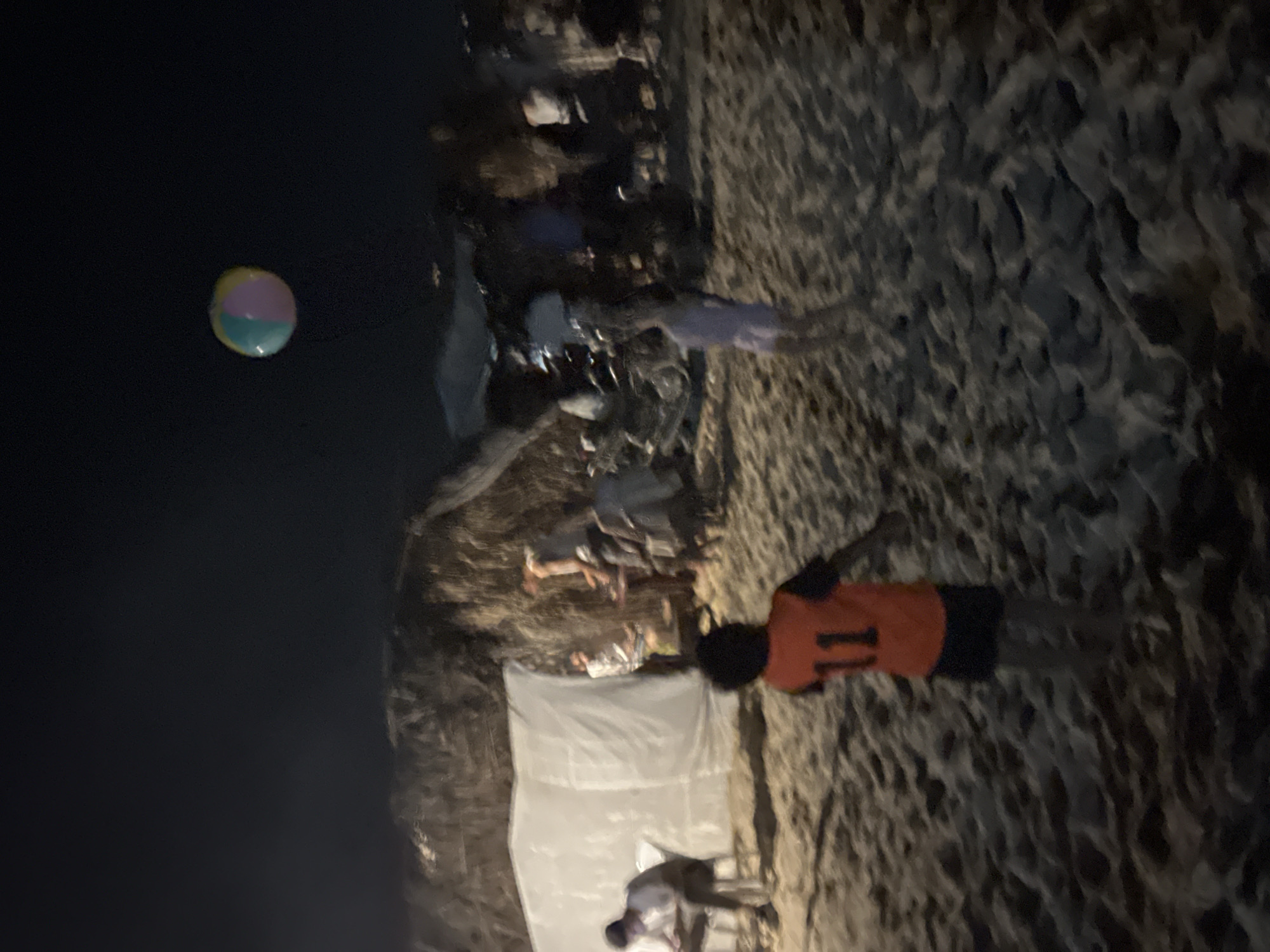 A nighttime aerial view shows a beach scene in Gaza, with a colorful beach ball suspended in the dark sky. In the foreground, a child wearing an orange vest with the number "11" and dark shorts stands on the sandy ground. Other figures and structures, including a white tent and a thatched-roof shelter, are visible in the background.