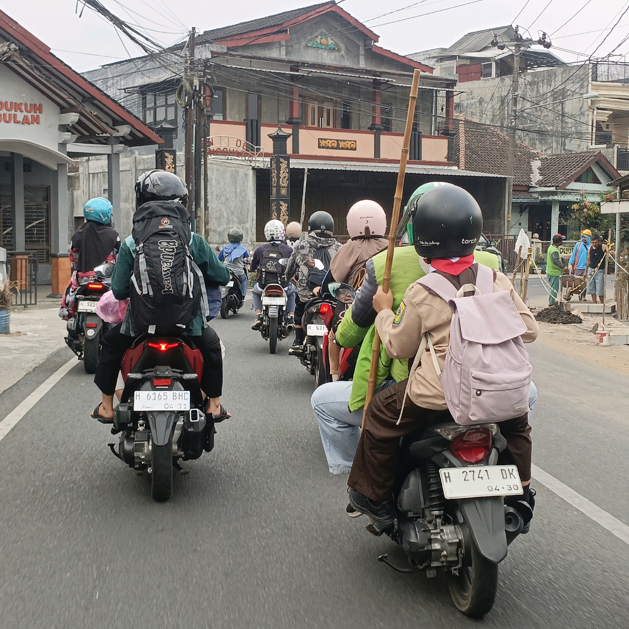 In Salatiga, Indonesia, a group of people are riding motorcycles on a street. The image focuses on the back of several motorcycles, with the riders wearing helmets and backpacks. One rider has a large black backpack with the word "kawanda" printed on it. Another rider is wearing a light pink backpack and a scouting uniform with a red scarf. Buildings line the street, some with red roofs and traditional Indonesian architecture. There are also utility poles with many wires overhead, indicating an urban or suburban setting. The road appears to be paved, and there is some construction work happening on the side of the road in the background, with men digging and piles of dirt. The sky is overcast. The license plates of the motorcycles are visible, with one reading "H 6165 BHC" and another "H 2741 DK".