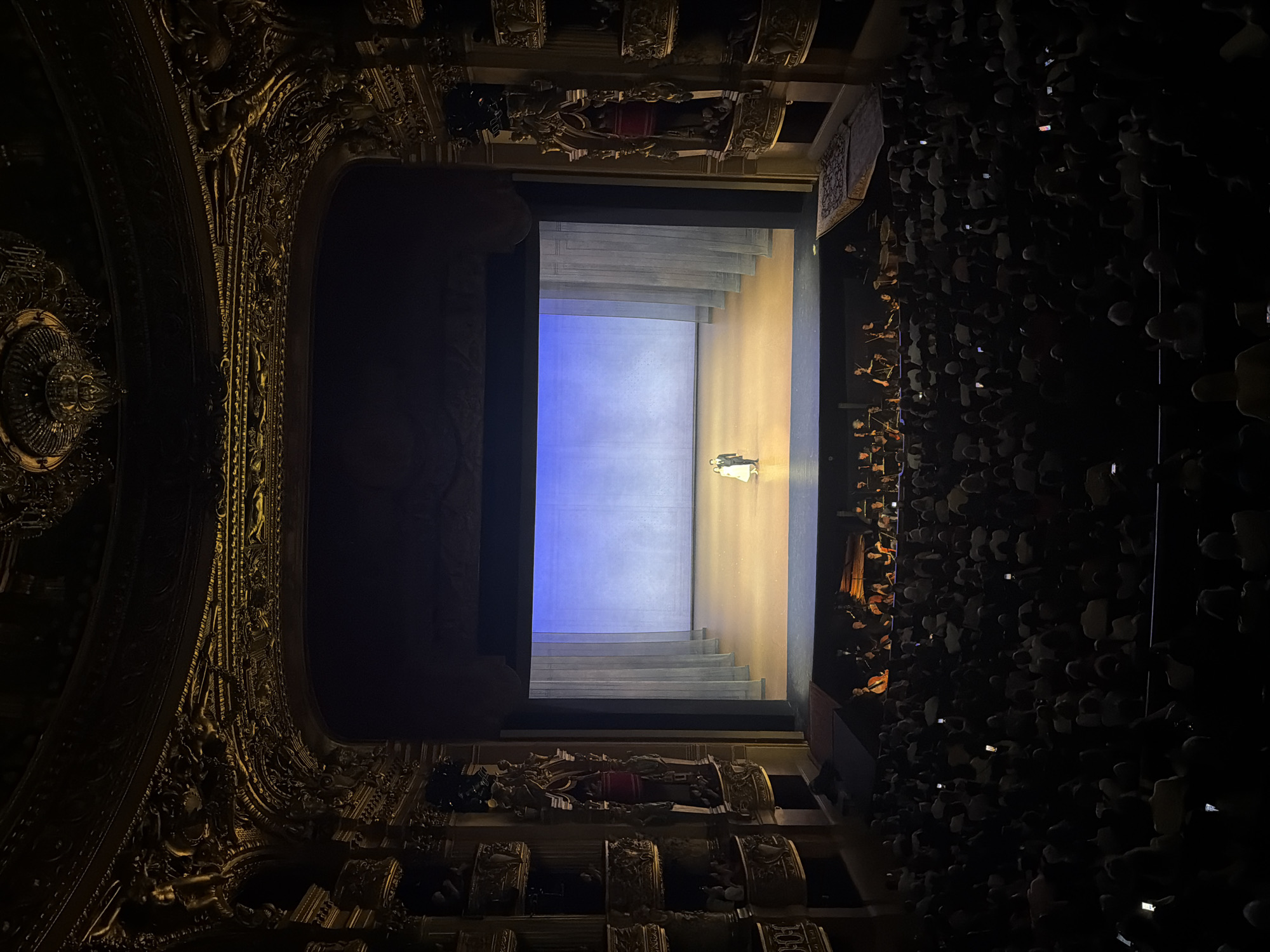 The image shows a view of the stage from an upper level box in the Palais Garnier opera house in Paris, France. A single figure in a light-colored garment stands on a stage with a blue backdrop and tiered platforms on one side. The box is elaborately decorated with gold detailing and sculptural elements. A large crowd of people fills the space to the right of the box. The view is from an elevated position, looking down towards the stage and the audience.
