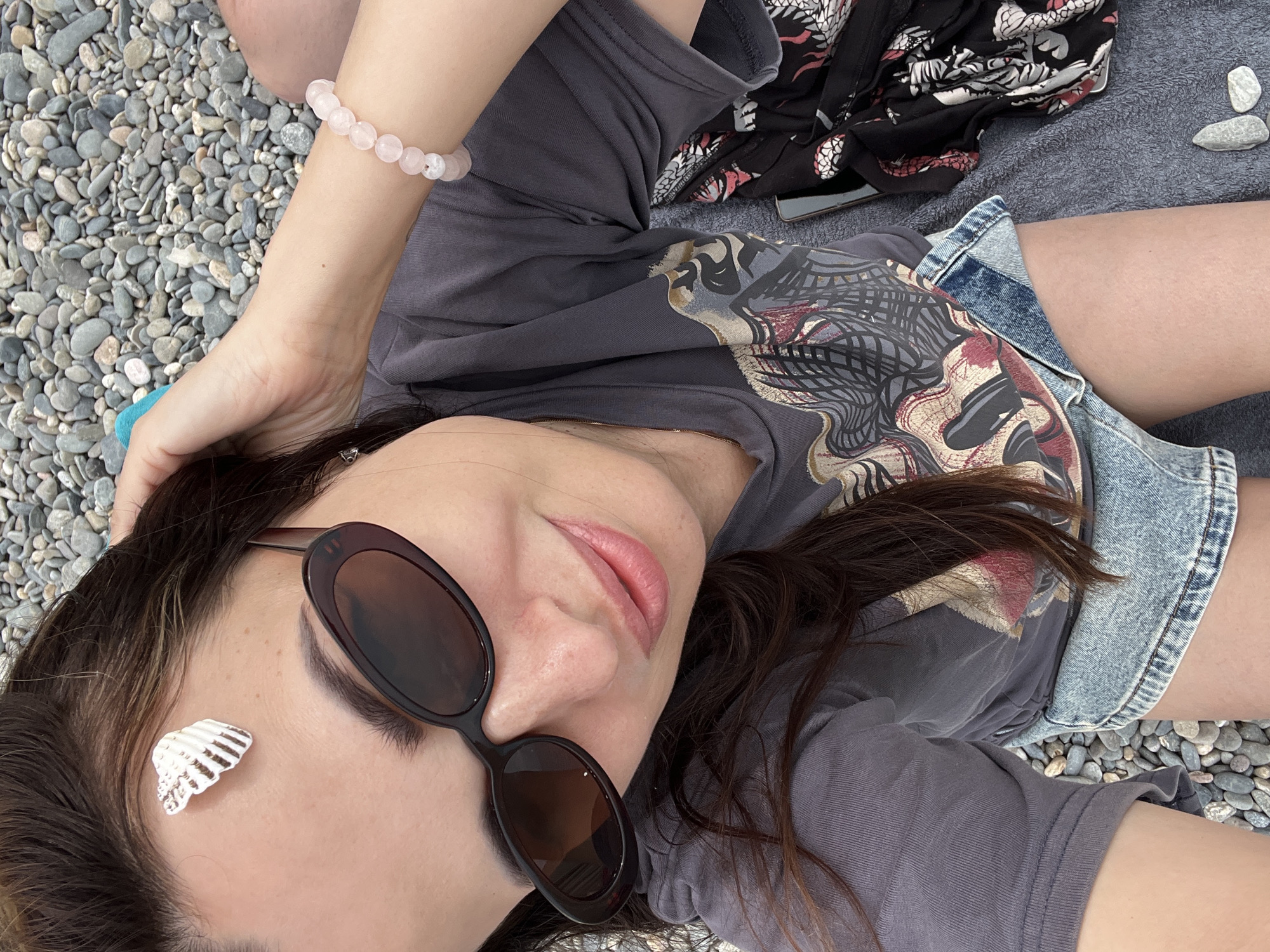 A woman sits on a pebble beach, looking at the camera. She is wearing sunglasses, a grey t-shirt with a graphic print, and denim shorts. A small seashell is stuck to her forehead. She has a pink beaded bracelet on her wrist. A black patterned garment and a mobile phone lie next to her. The background consists of grey and brown pebbles and the sea.