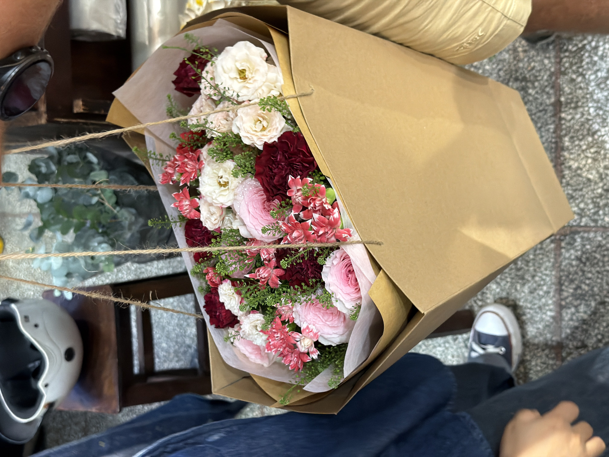 Here is a description of the image:
A high-angle shot shows a person holding a bouquet of flowers in a brown paper bag. The flowers are a mix of white, pink, and burgundy varieties. The person is wearing blue jeans and has one foot visible, wearing a navy and white sneaker. To the left of the flowers, out of focus, are sunglasses, a white helmet, and what appears to be greenery on a brown wooden table. The floor is patterned with gray tiles. The exact location in Nha Trang Ward, Vietnam is not discernable from the image.