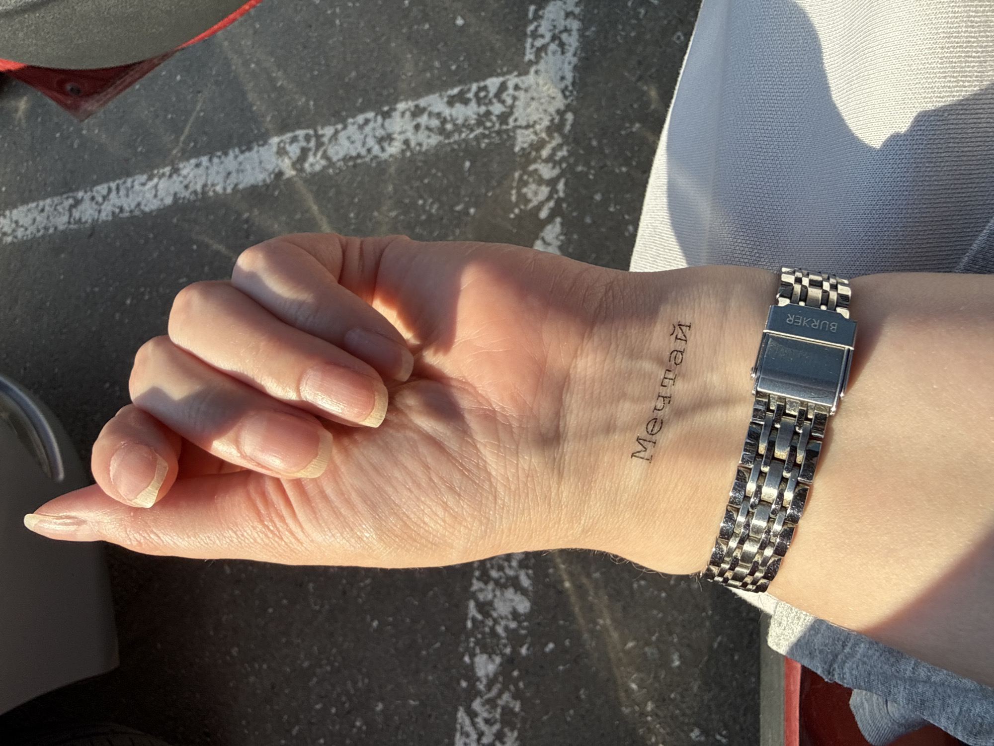 A person's left hand and wrist, adorned with a silver watch, are prominently featured in an outdoor setting. The hand is extended, palm-up, displaying a tattoo of the word "мечтай" (mechtay), which translates to "dream" in Russian. The wrist is encircled by a silver-colored watch with a deployant clasp. The surroundings consist of asphalt, marked with a white line, and glimpses of clothing in light gray and red hues. The location can be inferred to be Izhevsk, Russia, based on the Cyrillic inscription of the tattoo.