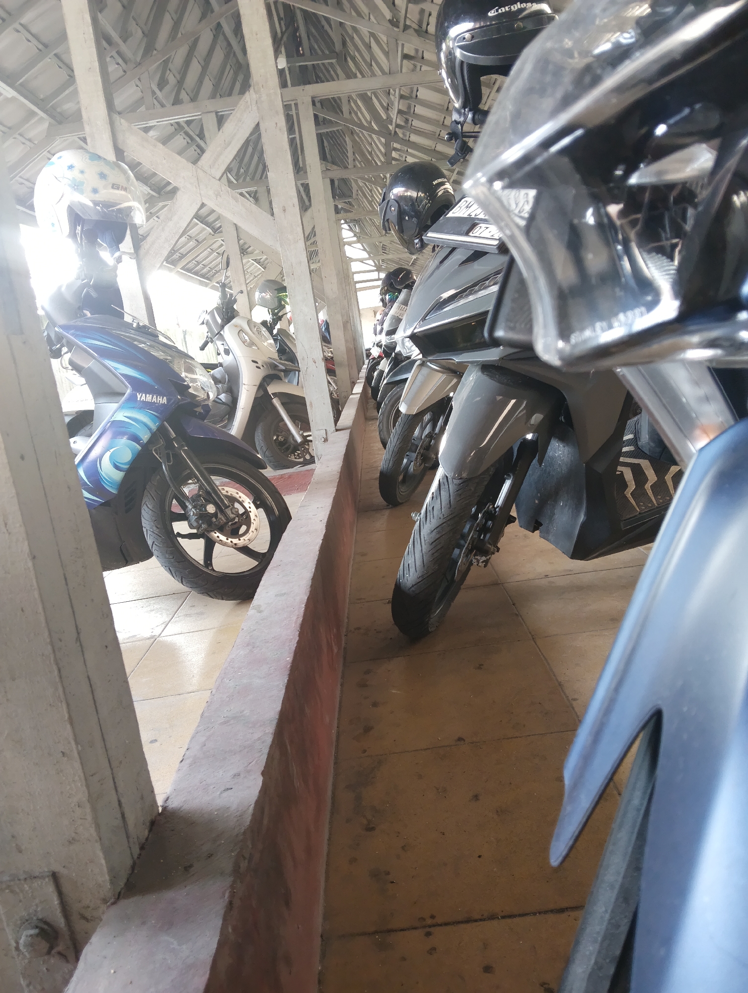 A line of parked motorcycles is visible under a covered area, likely a parking structure, with a light-colored tiled floor. The motorcycles are predominantly dark-colored, with a few lighter shades visible. Some helmets are placed on the handlebars of the motorcycles. The structure's roof is supported by exposed beams and trusses, suggesting an open-air or partially enclosed space. The image appears to be taken from a low angle, focusing on the front parts of the motorcycles. The environment suggests an urban setting in Pekanbaru, Indonesia, possibly a public parking area or a dedicated motorcycle parking zone. There are no people visible in the image, and the scene is static, depicting parked vehicles.