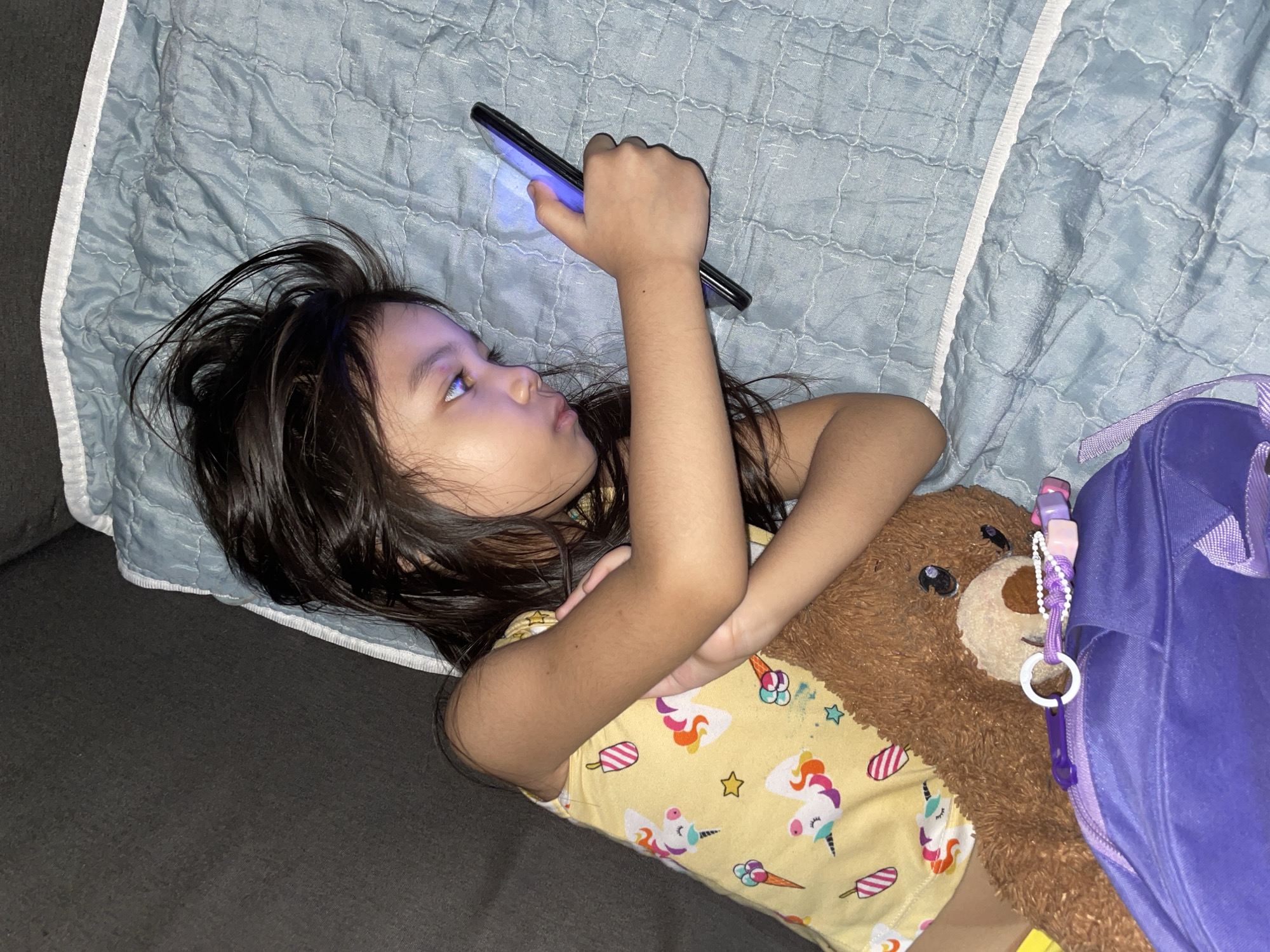 A young girl is lying on her back on a gray couch, propped up by a light blue quilted blanket. She is holding a black smartphone in her right hand, looking at it with her eyes wide open. Her dark hair is spread out around her head. She is wearing a yellow t-shirt with a pattern of unicorns and ice cream cones. Next to her, on her right side, is a brown teddy bear, and a portion of a purple backpack is visible. The lighting suggests it is either nighttime or a dimly lit room. The scene is casual and relaxed, with the girl appearing engaged with her device. The overall impression is one of a child enjoying screen time.