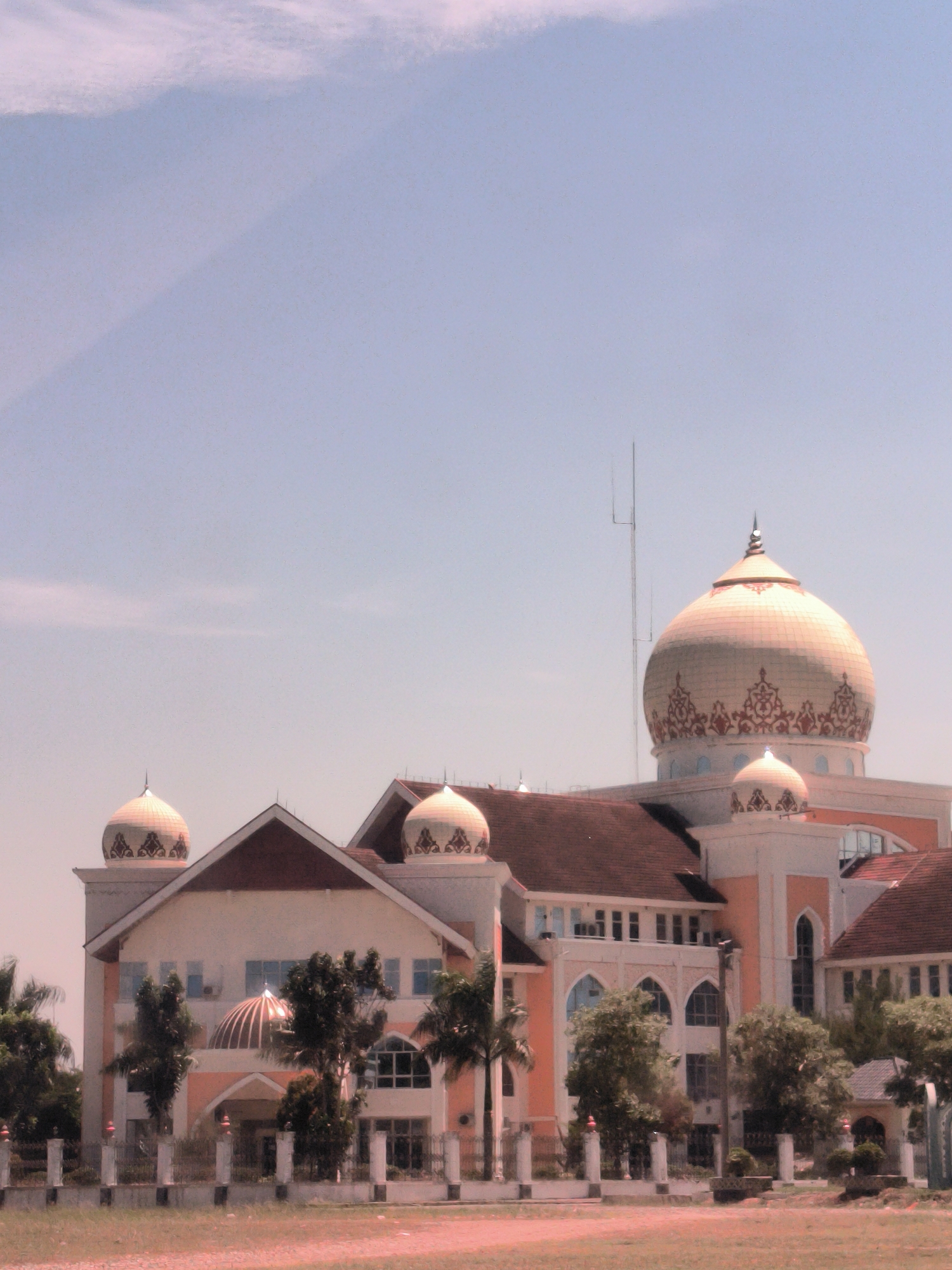 The image shows a large, ornate building with multiple domes located in Meunasahaluedrienlhoksukon Barat, Indonesia. The main dome is large and off-white with a decorative red and gold pattern around its base. Two smaller domes flank the main building. The building has a red-tiled roof and is painted in shades of white and peach. The architecture includes arched windows and decorative details. In front of the building, there is a fence and some trees. The sky is light blue with some faint cloud cover. A thin metal antenna is visible extending upwards from the roof of the building.