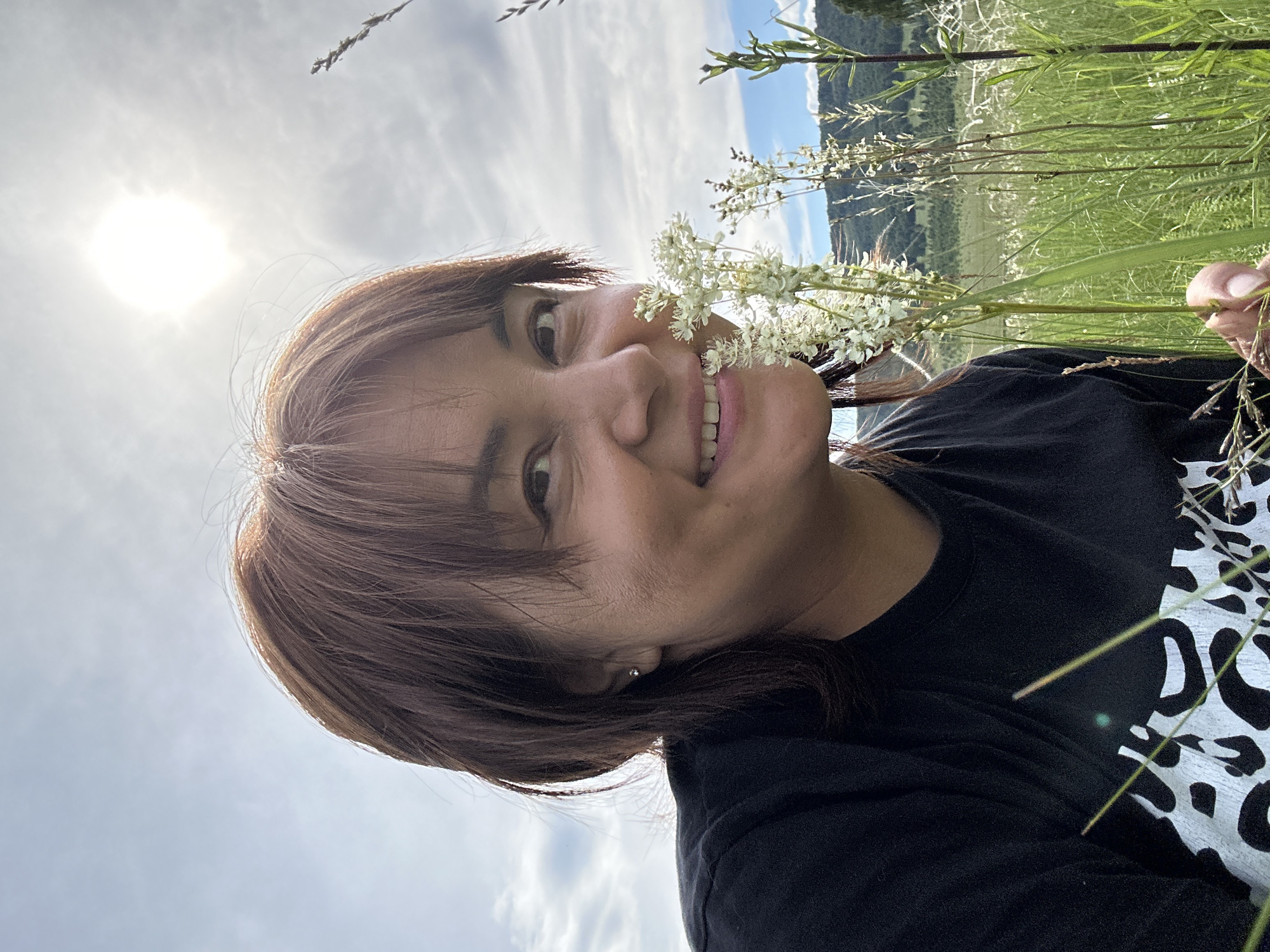 An outdoor, close-up, selfie-style photograph shows a woman with brown hair and fair skin smiling and holding a small cluster of white flowers near her face. She is wearing a black t-shirt with a white abstract pattern on the left side. The background features a field of tall green grass, some dry stalks of grass, and distant rolling hills under a bright, hazy sun and blue sky with scattered clouds. The location is Unknown, Unknown.