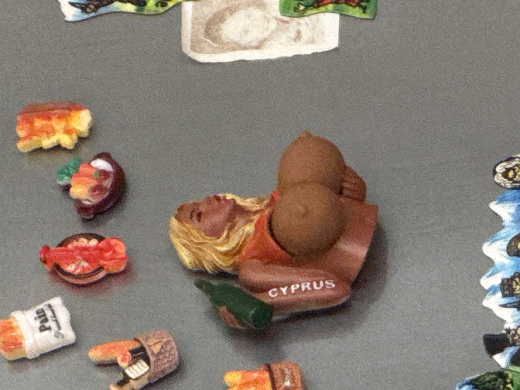 This is a close-up, eye-level, horizontal, medium shot of several refrigerator magnets adhered to a gray surface. In the center of the image is a magnet shaped like a busty blonde woman wearing an orange top. The word "CYPRUS" is printed vertically on her arm. She is holding a green bottle over her shoulder. To the left of the woman magnet, there are two other magnets. One is a white bag with "Pain" printed on it, and the other is a brown basket with breadsticks in it. Above the woman magnet, and to the left, are two small magnets, one shaped like a red shrimp and the other like a small bowl of food with fruit. To the far right of the woman magnet is a magnet with a black and white portrait of a child. At the bottom of the image, there is a magnet depicting a landscape with snowy mountains and buildings.