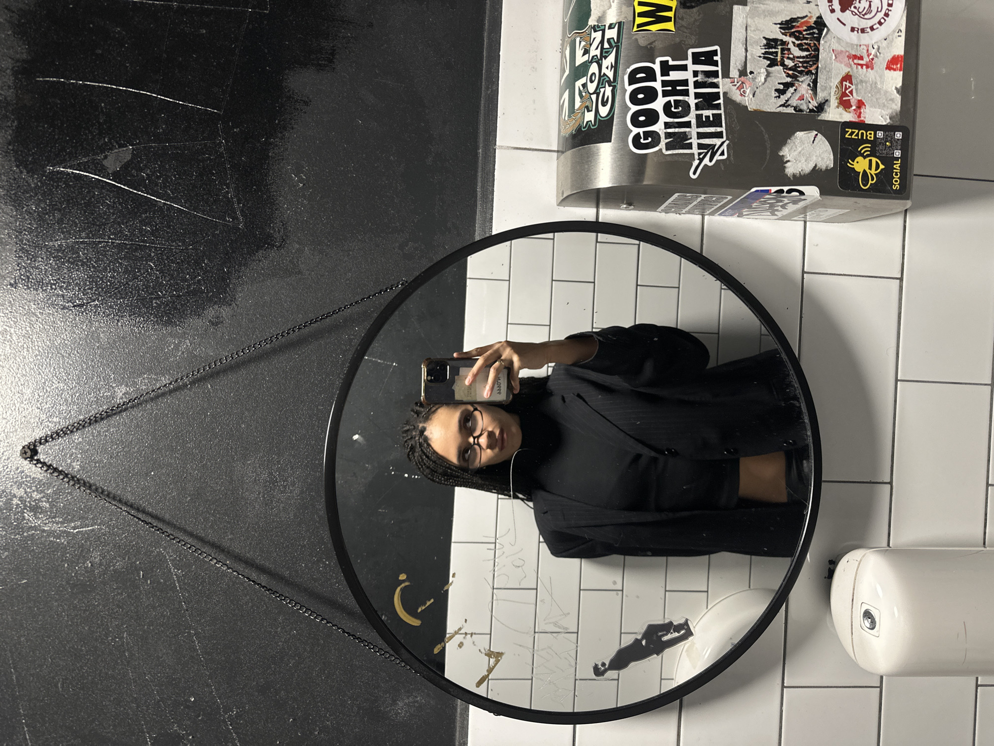 The image shows a mirror selfie taken by a person in what appears to be a public restroom or utility area, set in London, United Kingdom. A circular mirror with a black frame is centrally positioned, suspended by a black chain from a dark, textured wall on the left side of the frame.
Reflected in the mirror is a person with dark skin, braided hair, and glasses, wearing a dark turtleneck and a pinstriped blazer. She is holding a smartphone (visible in her hand) to capture her reflection, looking directly into the camera with a neutral expression.
The background visible in the reflection consists of white subway tiles, which also frame the mirror on its right side. On this tiled surface, some graffiti is visible, including what appears to be a golden crescent moon and other white scribbles. A black silhouette sticker, resembling a person, is also adhered to the mirrored surface.
Above the mirror, a grey dispenser or container is heavily adorned with various stickers. Prominently visible texts on these stickers include "GOOD NIGHT VIENNA," "BUZZ" next to a bee icon and a QR code, and "SOCIAL" also with a QR code. Other partially visible text includes "RECORDS." To the right of the mirror, a white, rounded hand dryer or similar device is mounted on the white tiled wall. The dark wall to the left of the mirror exhibits several white scratches and marks. The scene suggests an indoor, urban environment.