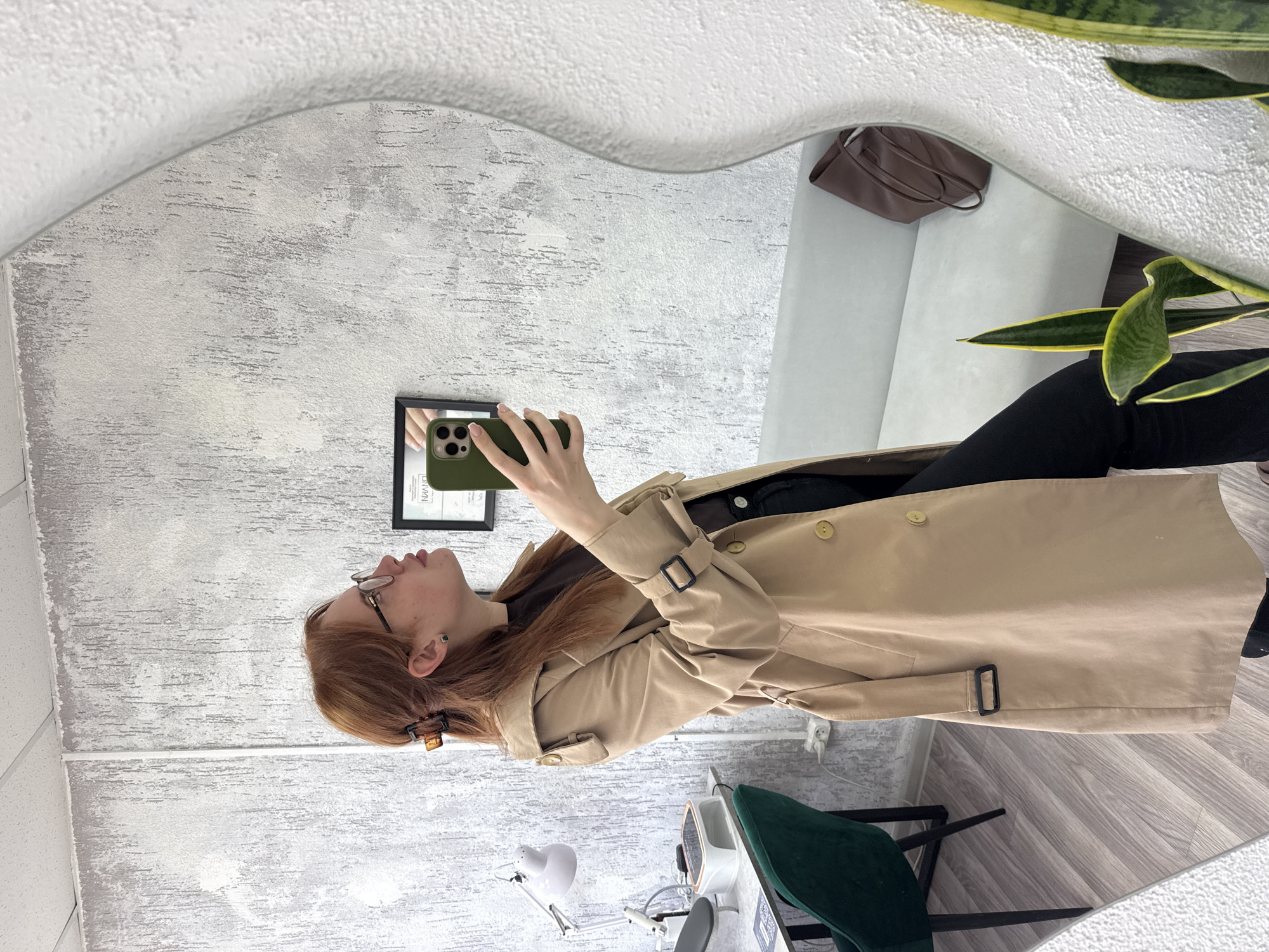 A young woman with reddish-brown hair and glasses is taking a mirror selfie in an indoor setting. She is wearing a light beige trench coat over dark clothing, with her hair partially held back by a tortoiseshell claw clip. She holds a smartphone with a green case, its multiple camera lenses visible as she photographs her reflection.
The mirror, which has a distinct wavy or heart-shaped top edge, reflects the woman and the area behind her. Visible in the reflection are a light grey sofa or bench, a brown handbag resting on it, and a tall snake plant with variegated green and yellow leaves.
Outside the mirror's reflection, to the right of the woman, a green velvet chair and a white desk with a flexible white desk lamp are visible, along with other equipment, suggesting a beauty or nail salon environment. The wall features a textured, light grey and white finish. The ceiling is constructed from white acoustic tiles, and the floor has a light wood-look laminate.
A framed document or picture, partially visible on the wall both directly and in the mirror, shows the text "URWAN". The phone screen, also visible in the mirror's reflection, displays faint graphical elements that might be part of a calendar application. The scene captures a casual moment in what appears to be a modern commercial space.