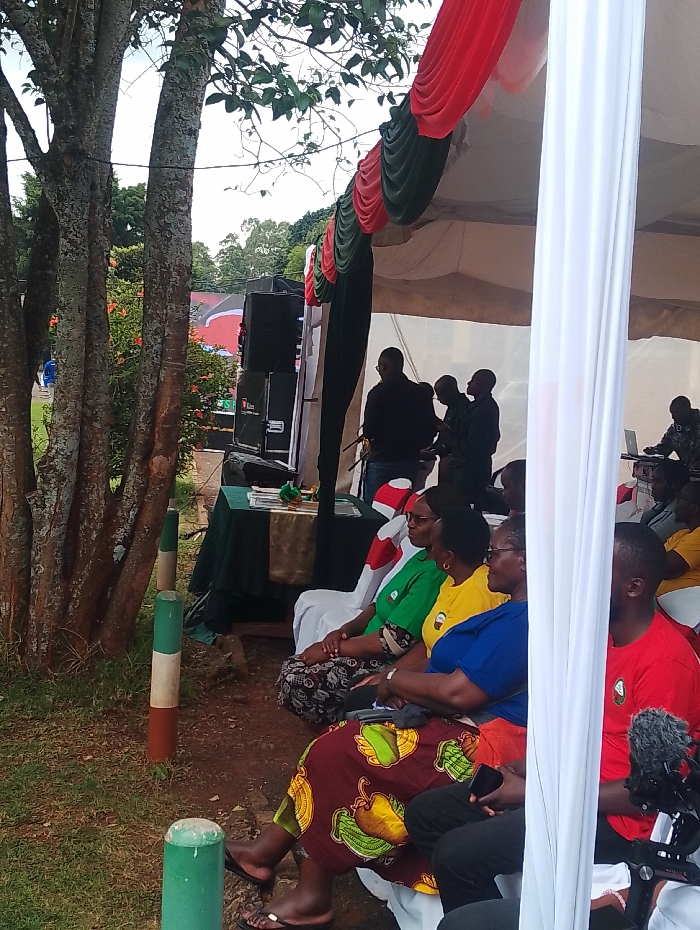 An outdoor gathering is depicted under a large white tent, adorned with drapes in red and dark green, colors reminiscent of the Kenyan flag. The setting appears to be a natural, somewhat rustic environment, possibly a park or open ground in Gitaru, Kenya.
In the foreground to the left, several large trees with textured bark and leafy canopies are prominent. At their base, green and brown/red painted bollards further echo the national colors.
Under the tent, a group of attendees, primarily adults, are seated on white plastic chairs, facing an area with sound equipment. Several women in the foreground wear bright, solid-colored t-shirts (green, yellow, blue) and a traditional patterned skirt in maroon and green hues, suggesting diverse attire. Their posture indicates they are attentively observing an ongoing event. Further back on the right, a man in a red t-shirt is partially visible, also seated.
Towards the center of the tent, several men are standing near stacked sound speakers and a green-covered table. One man appears to be at a microphone or speaking into a device, while another is seated at a table further back, working on a laptop. A professional microphone setup is visible in the bottom right corner, indicating that the event is being recorded or amplified.
The lighting suggests it is daytime, likely overcast, as there are no harsh shadows. The ground under the tent is dirt, transitioning to grass on the left. The overall scene conveys an organized yet informal public or community event, possibly a speech, presentation, or cultural performance. No legible text is clearly visible in the image.