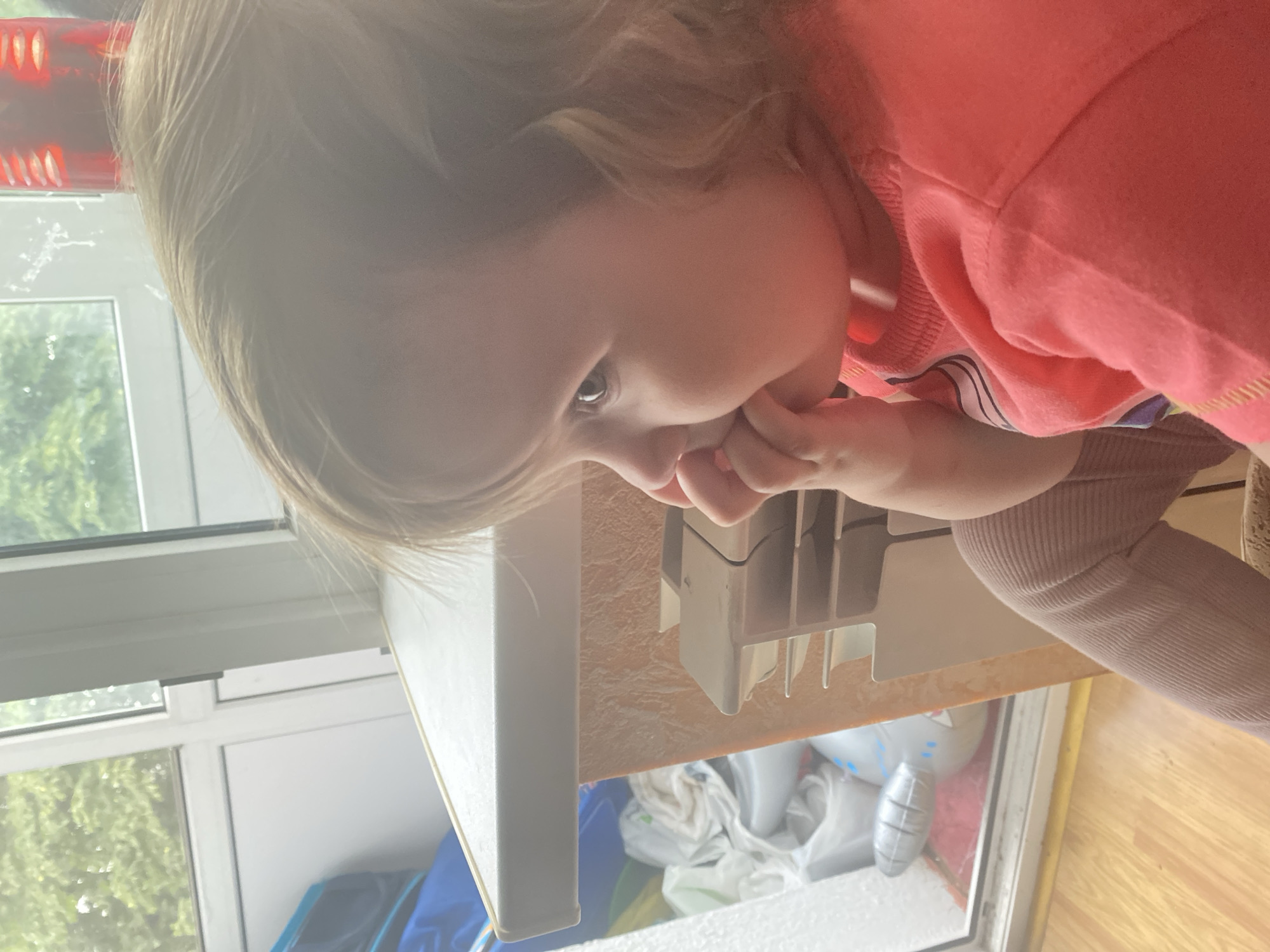 A young child is pictured in a close-up, profile view, with their right hand to their mouth. The child is wearing a coral-colored shirt and light brown leggings. They are seated near a window, and through the window, greenery can be seen. A table or counter is positioned in front of the child, with some items on it and a heater-like object attached to its side. The floor is made of wood.