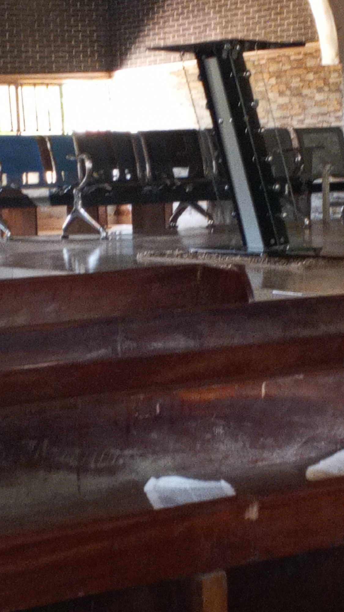 The image depicts an indoor waiting area, possibly an airport or bus station, with rows of dark seats. A metal structure with cables runs vertically in the background. The floor appears to be a polished surface, reflecting the light. In the foreground, a dark wooden bench with white paper scraps on it is visible. The overall lighting suggests an overcast day or an indoor setting with artificial lights. The scene is devoid of any people. The specific location is Unknown, Unknown.