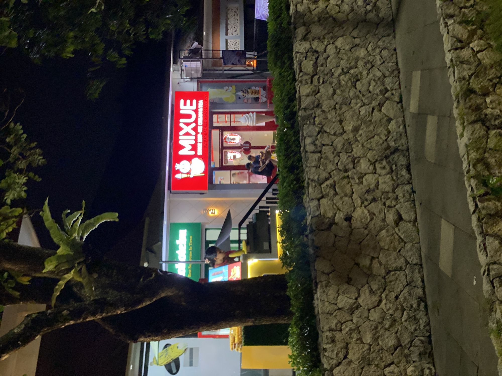 In Kuta Selatan, Indonesia, at night, a street view shows a Mixue ice cream and tea shop. The shop's bright red sign with "MIXUE" in white letters is illuminated. To the left of the shop, a green sign with white text reads "ohotoboo" and "Best Supreme In Town". Inside the Mixue shop, a person with dark hair is visible behind a counter, and two customers are seated. Ice cream cones are displayed outside the shop. The shop is situated in front of a stone wall, with a paved walkway in front of the wall. Lush green bushes are planted along the top of the stone wall. A large tree with ferns growing on its trunk stands to the left of the frame, obscuring part of the view. The surrounding area is dark, suggesting it is nighttime.