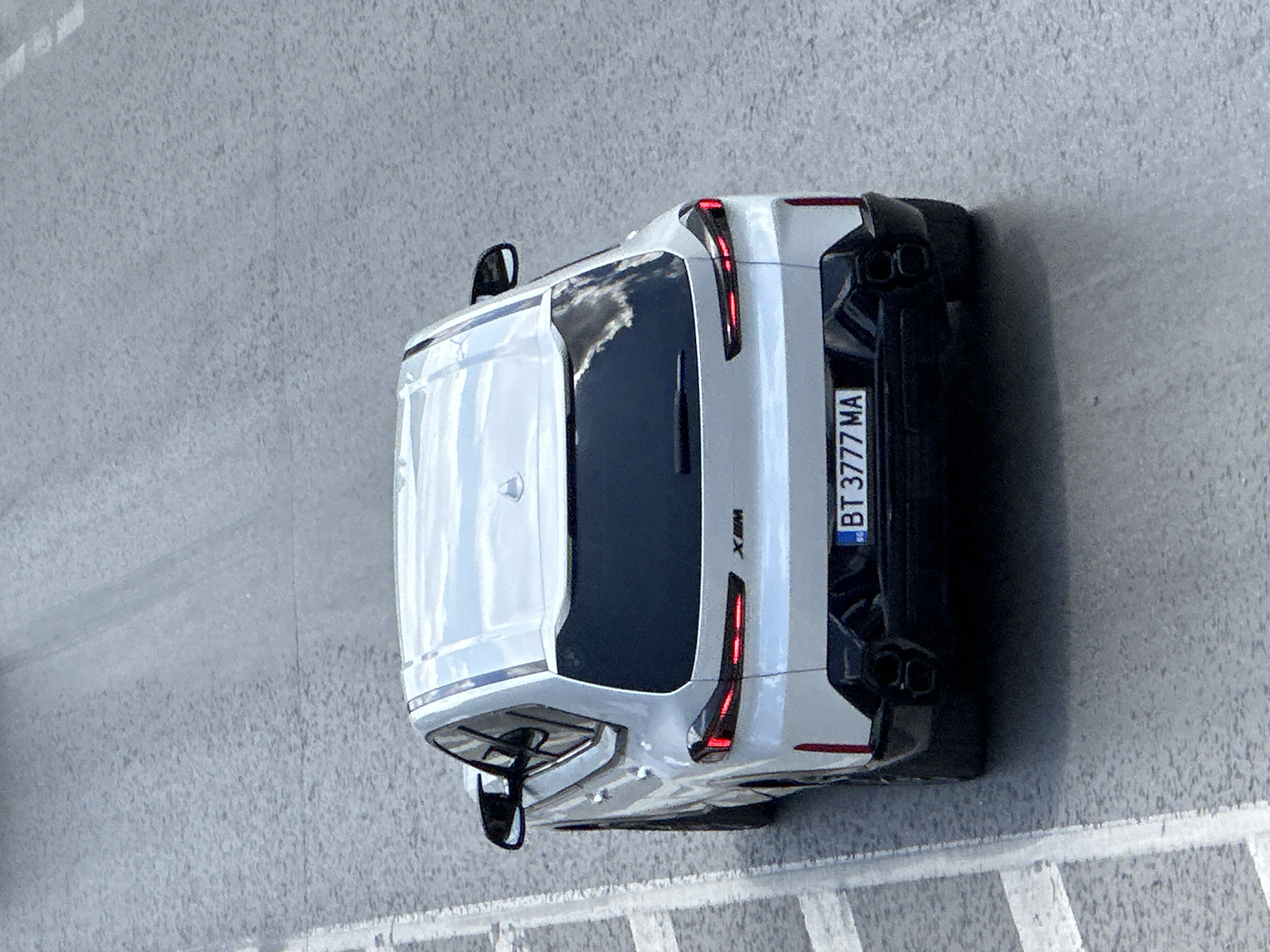 A white BMW XM is parked on a paved road, likely in Veliko Tarnovo, Bulgaria, given the license plate format. The car is viewed from the rear at an elevated angle. The vehicle has black accents on the rear bumper, including quad exhaust pipes. The license plate reads "BT 3777 MA". The road surface appears to be asphalt with visible white lane markings on the left side of the frame. The lighting suggests it is daytime, with reflections visible on the car's windows and roof. There are no people or other vehicles immediately present in the frame.