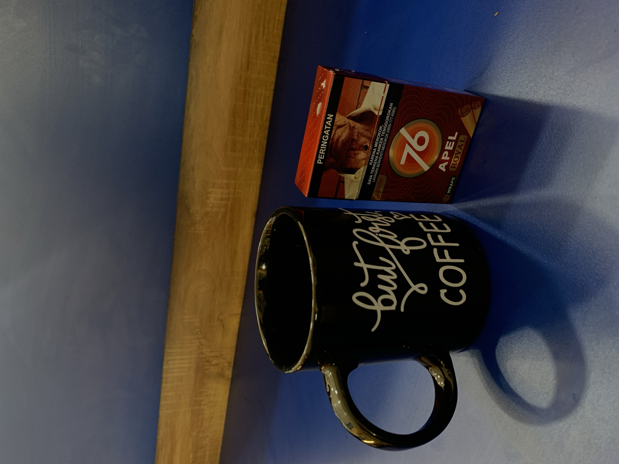 A close-up, eye-level shot captures a black mug with white cursive text that reads "but first coffee." The mug is positioned on a blue surface. To the right of the mug is a red and brown cigarette pack labeled "76 APEL ROYAL." The pack has an image of a man's face and text in Indonesian. The background features a textured wooden beam against a dark blue wall. The scene appears to be indoors, possibly in a cafe or home environment. The lighting suggests a single light source casting a shadow from the mug. There are no individuals present. The image does not provide any specific clues about the location within the city of Padangjaya, Indonesia, beyond the general presence of a flat blue surface, a wooden beam, and a blue wall.