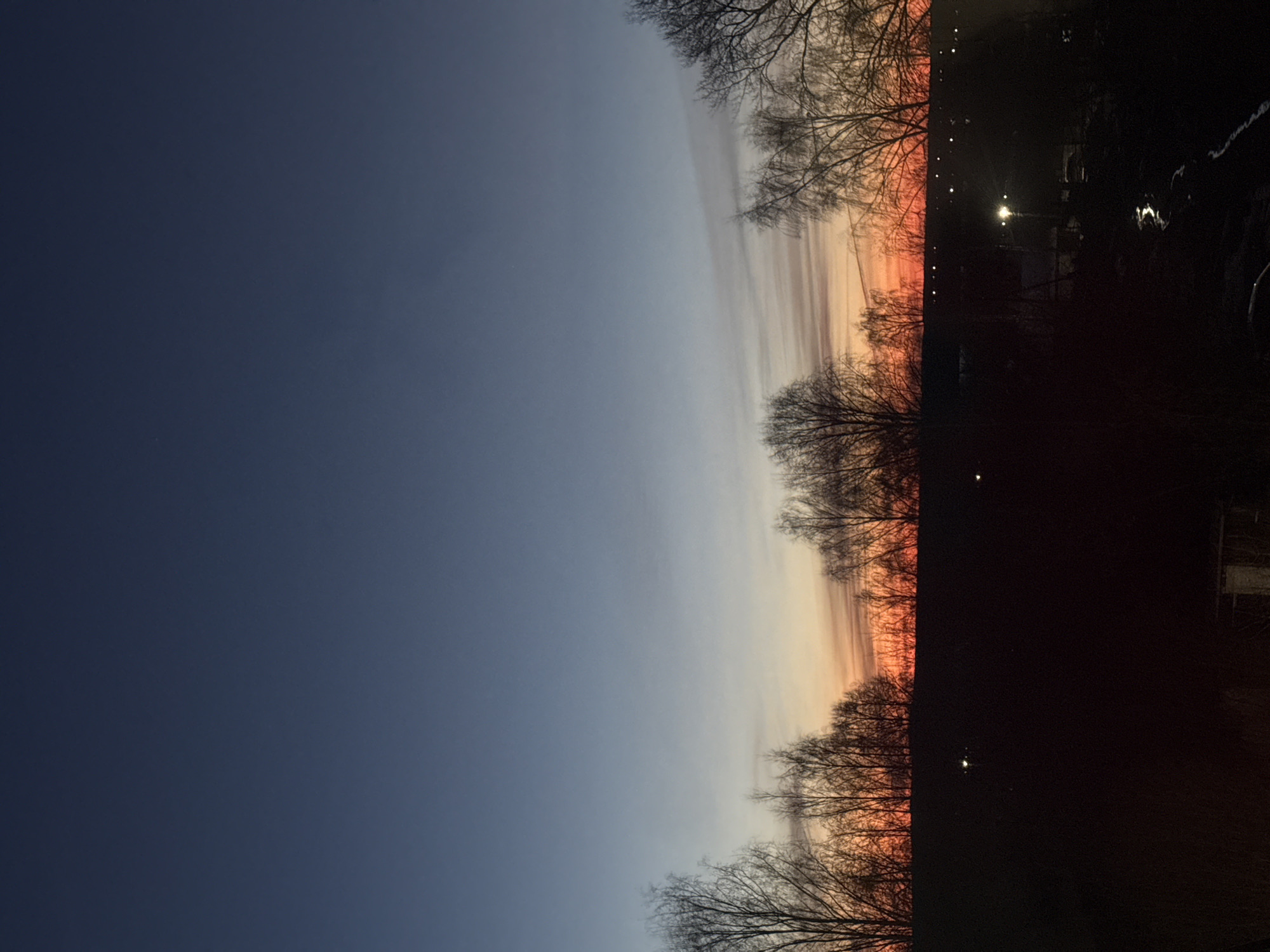 The image shows a sky at dusk or dawn. The sky transitions from a dark blue on the left to orange and yellow hues on the right, suggesting the presence of the sun below the horizon. Silhouetted trees are visible along the bottom and upper edges of the colorful sky. The right side of the image is dark, with the faint outlines of buildings and a lit street lamp. There is no indication that the location is in Chaikovsky, Russia. The image doesn’t contain any people or any actions.