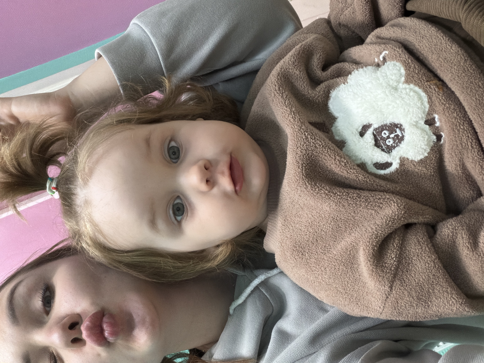 The image shows a selfie taken by an adult female and a young child. The child, lying on the female's chest, is fair-skinned with light brown hair tied in a ponytail with a colorful hair tie. The child is wearing a brown sweater with a white bear applique. The adult female is fair-skinned with dark eyebrows and full lips puckered in a kiss. Both are lying on a light gray fabric with a pink and mint-colored mat in the background. The image was taken in Chaikovsky, Russia.