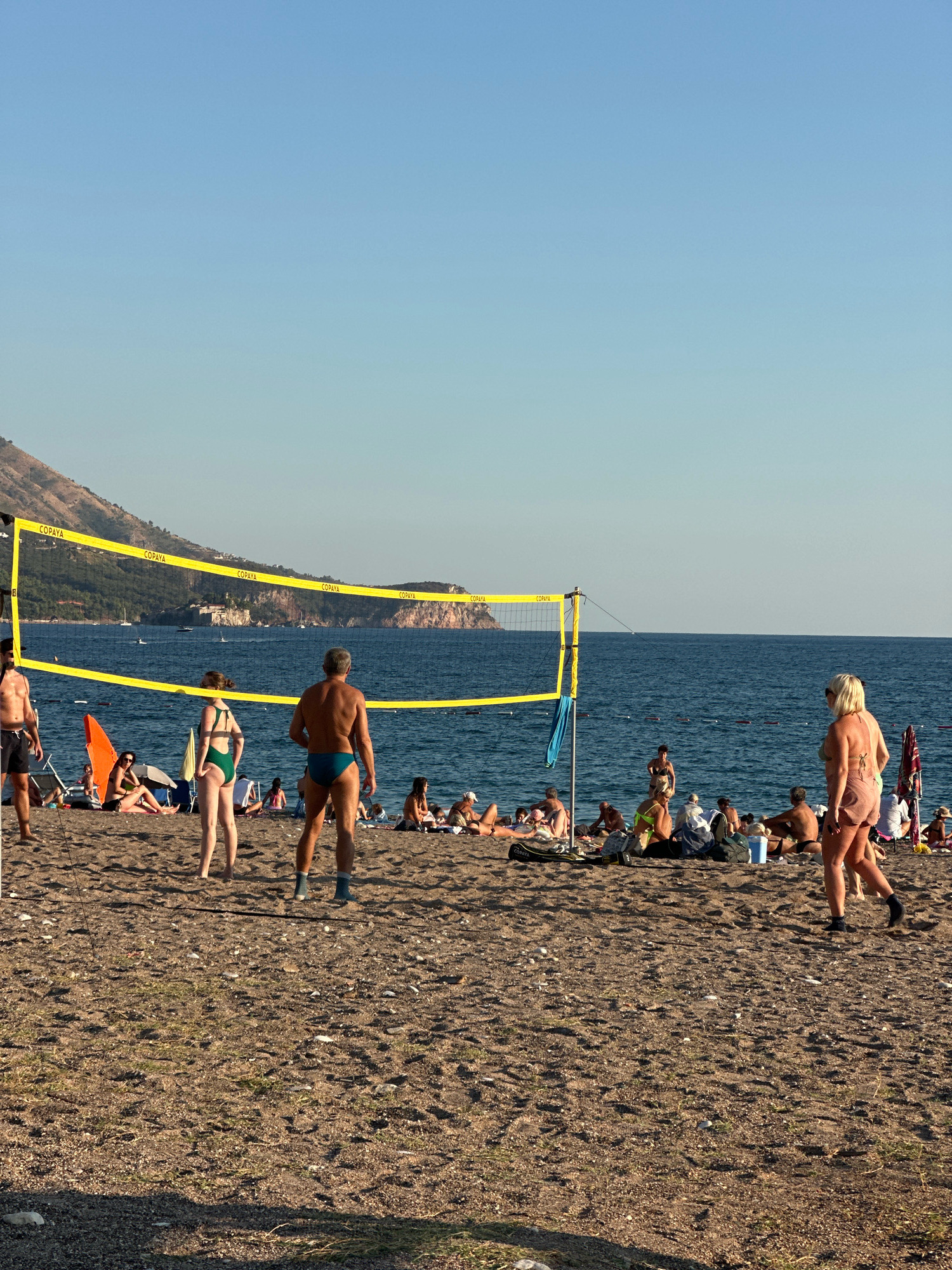 The image depicts a beach scene in Boreti, Montenegro, on a clear day with a blue sky. A volleyball net with yellow boundaries stretches across the sand, indicating a game in progress or set up for play. Several people are visible on the beach. In the foreground, a man with grey hair and a blue swimsuit stands facing away from the camera, with his back to the viewer. To his left, a woman in a green bikini is also positioned facing away. Further left, a man in dark swim trunks stands with his back to the viewer. In the mid-ground, numerous individuals are scattered across the beach, some sunbathing, others sitting and conversing. The sea stretches out beyond the beach, with several boats visible in the distance. Mountains form the backdrop, with vegetation covering their slopes. The sand is a mixture of pebbles and fine particles. The overall atmosphere is one of leisure and recreation on a sunny day.
