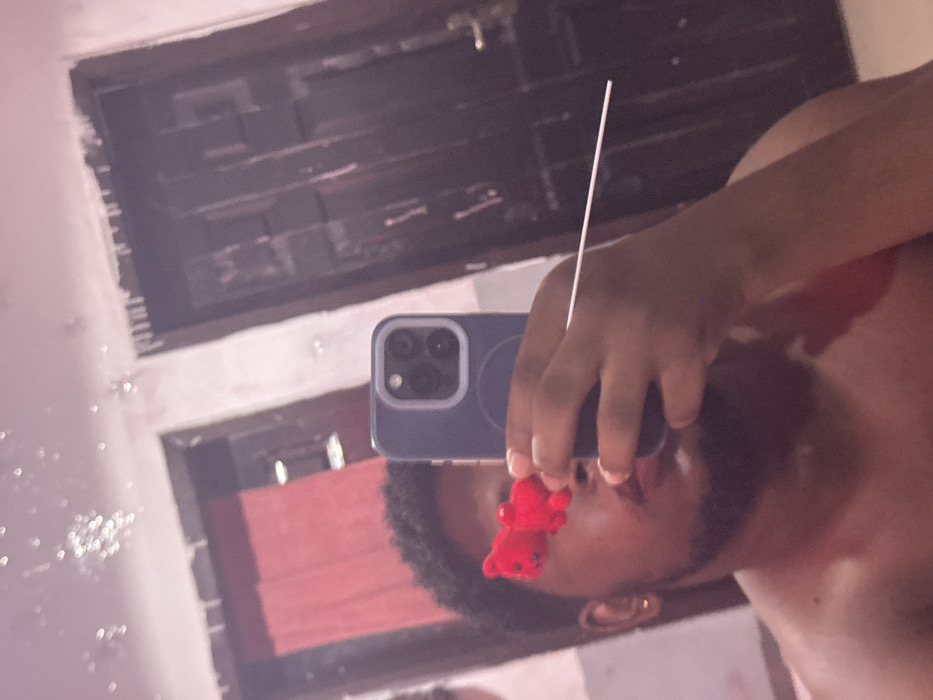 The image captures a mirror selfie of a male individual indoors in Ipetumodu, Nigeria. The subject, visible from the chest up, has dark, short, curly hair and a beard, and wears a small gold stud earring in his left ear. He appears shirtless. He holds a blue smartphone, likely an iPhone, in his right hand, partially obscuring his face. A small, red, two-part decorative object is affixed to the phone, positioned over his eyes in the reflection. A thin, white, horizontal rod extends from the right side, also seemingly held by his right hand. The mirror surface shows several light specks or imperfections. The reflected background reveals interior details: a reddish curtain visible through an opening on the left, and a dark, paneled wooden door with a handle and lock on the right. The walls are light-colored, exhibiting some discoloration and minor surface irregularities, particularly near the top.