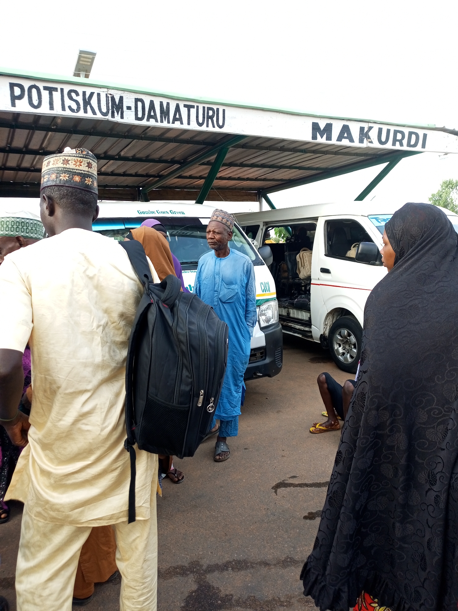 In the city of Gombe, Nigeria, at a transportation hub, a group of people are gathered near white minibusses. Overhead, a sign reads "POTISKUM-DAMATURU MAKURDI" in black capital letters. The ground is paved with asphalt, and there is a covered waiting area supported by poles. Several individuals, dressed in traditional Nigerian attire, are visible. One man in the foreground is wearing a light-colored, long-sleeved garment and a patterned cap, with a black backpack on his back. Another man, wearing a blue kaftan and a cap, stands to the right. A woman in a black hijab is also present, facing away from the camera. Inside the open side doors of a minibus, luggage can be seen. The scene suggests a departure or arrival point for inter-city travel.