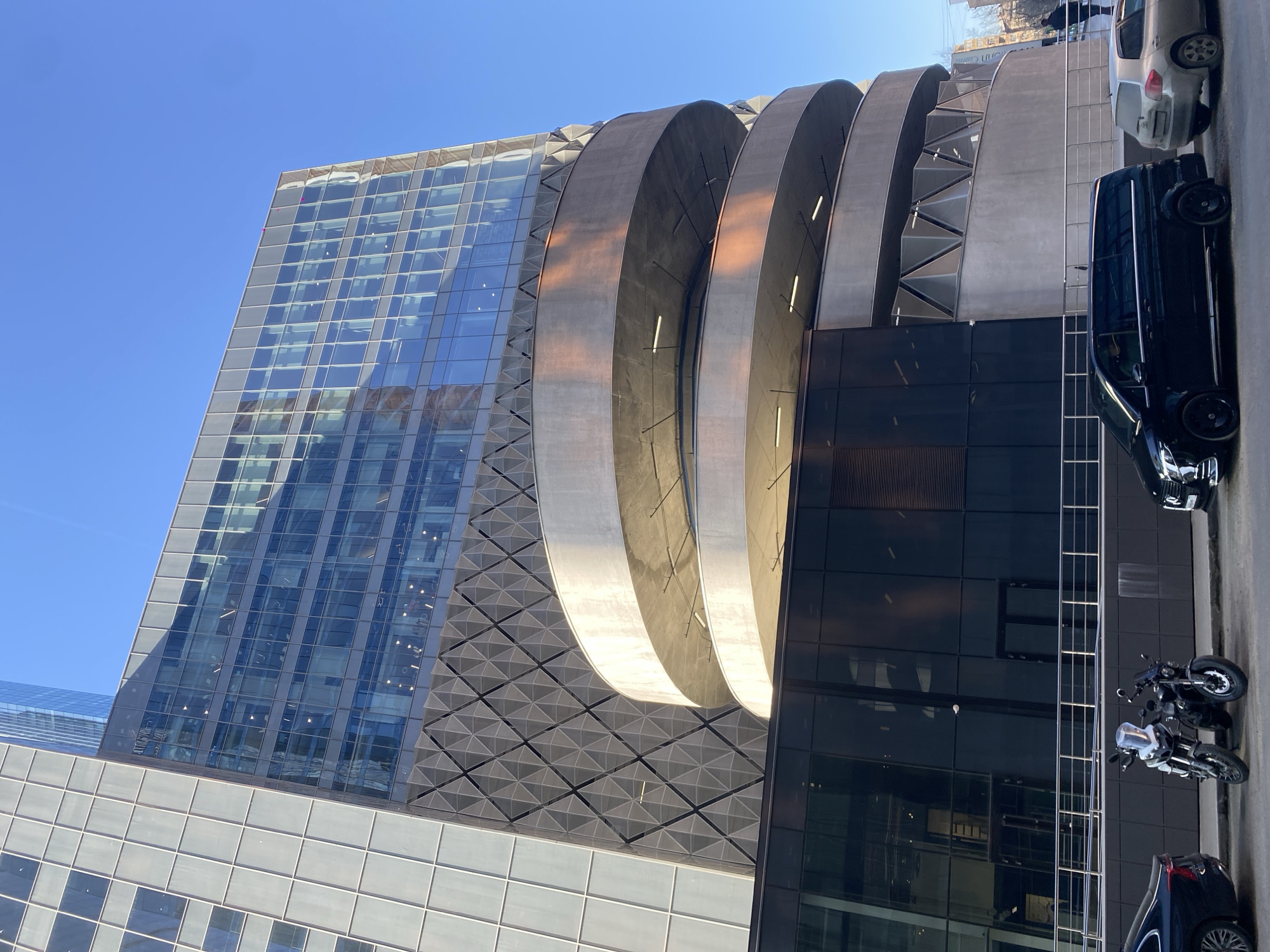 The image depicts a modern architectural structure in what appears to be an urban environment. A tall, glass-facade building is a prominent feature, reflecting the clear blue sky. Adjacent to this building are three large, curved concrete elements, possibly part of the building's design or a separate structure attached to it. Below the concrete elements is a section of dark, possibly black, glass.
Parked along the side of the building, likely on a street, are several vehicles. A black van is visible, and a motorcycle is parked next to it.
Based on the instruction to include the location in Khimki, Russia, it can be inferred that this architectural structure is located in Khimki, Russia. The specific architectural style is contemporary and the environment is indicative of a modern urban space with a combination of glass, concrete, and vehicular traffic.