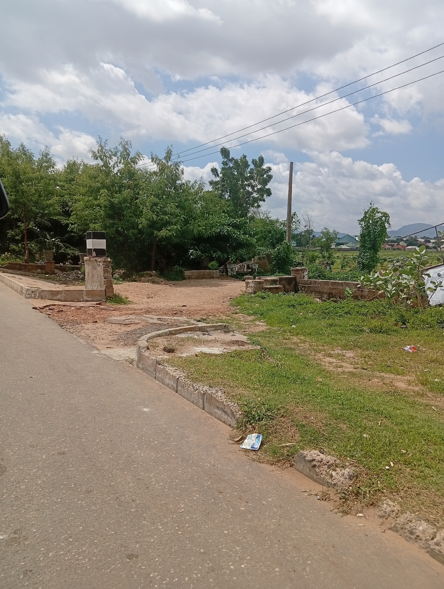 The image depicts an outdoor scene near a road in Jalingo, Nigeria. In the foreground, a paved road with some visible debris runs from the lower left towards the upper right. The road is bordered by a concrete curb, which is partially broken in places. Beyond the curb, there is an area of dry, sandy soil with patches of green grass.
In the mid-ground, several structures are visible. A concrete pillar with a black and white sign is situated on the left. To the right of the pillar, there are low, broken brick structures and more sandy ground. Further back, a line of lush green trees forms a backdrop.
In the background, a distant landscape includes more vegetation, buildings with green roofs, and hills under a cloudy sky. There are also power lines stretching across the sky. The overall impression is of an undeveloped or partially developed area on the outskirts of a city. No people are visible in the image.