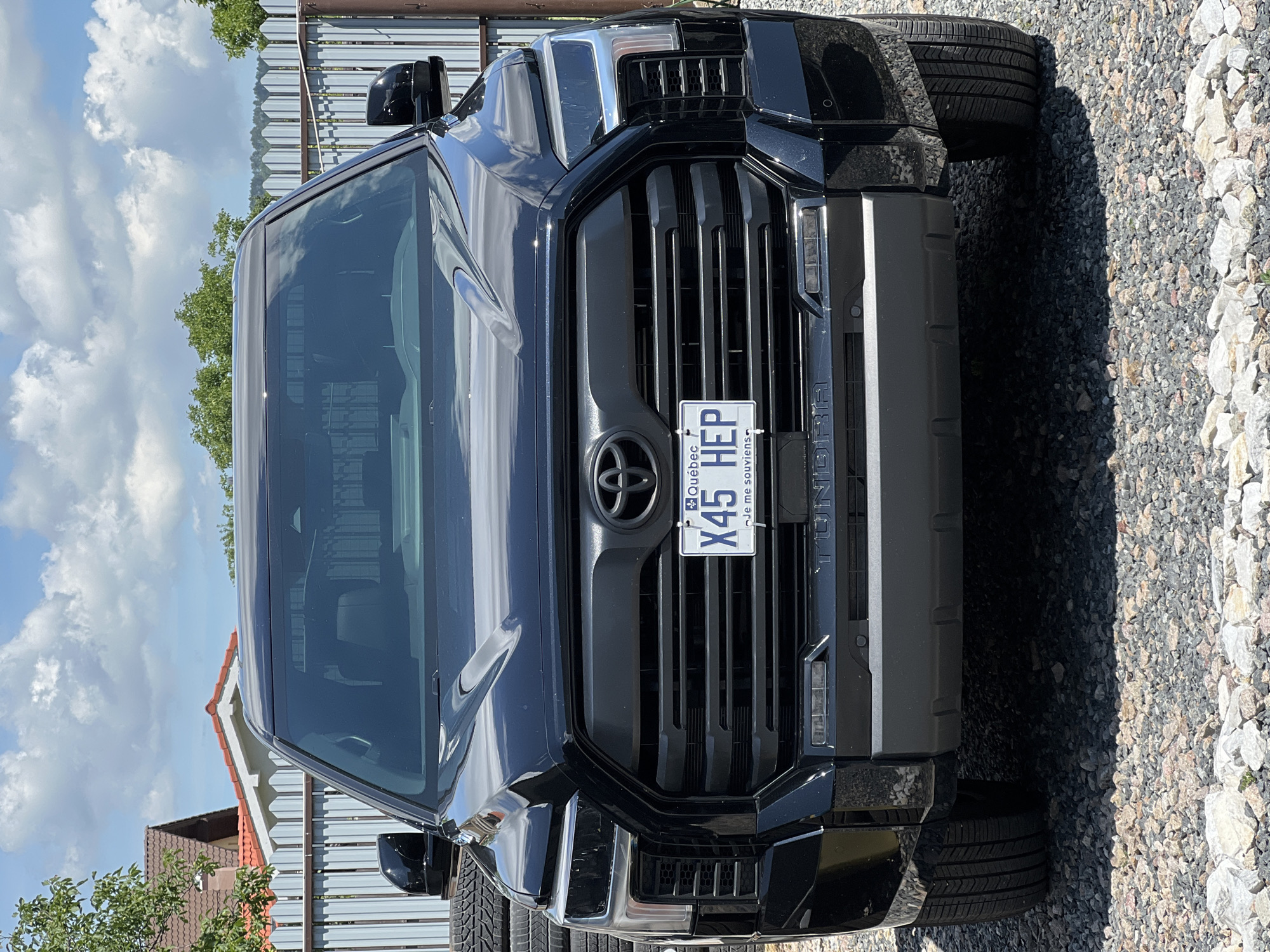 An outdoor, eye-level, full-shot photograph shows the front of a black Toyota Tundra pickup truck. The truck is parked on a surface of gravel and stones. Behind the truck is a gray metal fence and a residential building with a red-tiled roof. The truck's license plate reads "X45 HEP." The truck is clean and in good condition. The sky is blue with white clouds. The scene is well-lit by natural sunlight. There is no indication of the location being Cluj-Napoca, Romania.
