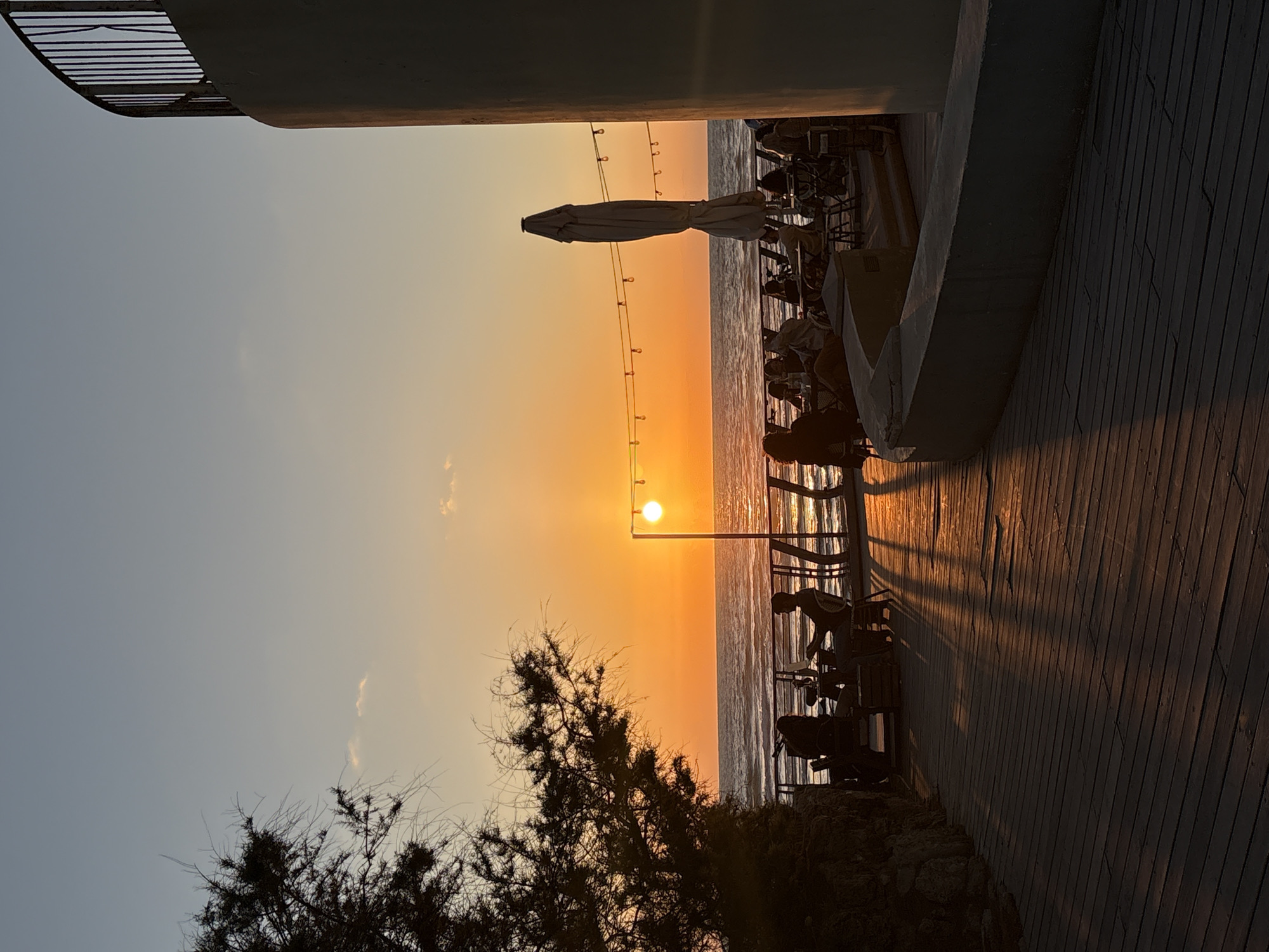 The image depicts a sunset scene over the Mediterranean Sea in Tel Aviv, Israel. The sun is setting directly over the water, creating a bright orange glow on the horizon and reflecting on the water's surface. Silhouetted figures of people are seated at tables on an elevated boardwalk area. A closed white umbrella and strings of lights are also visible. A tree with dark foliage is in the foreground on the left. The boardwalk extends horizontally into the frame, showing the textured wooden planks. Architectural details, including a curved wall, frame the scene on the upper right.