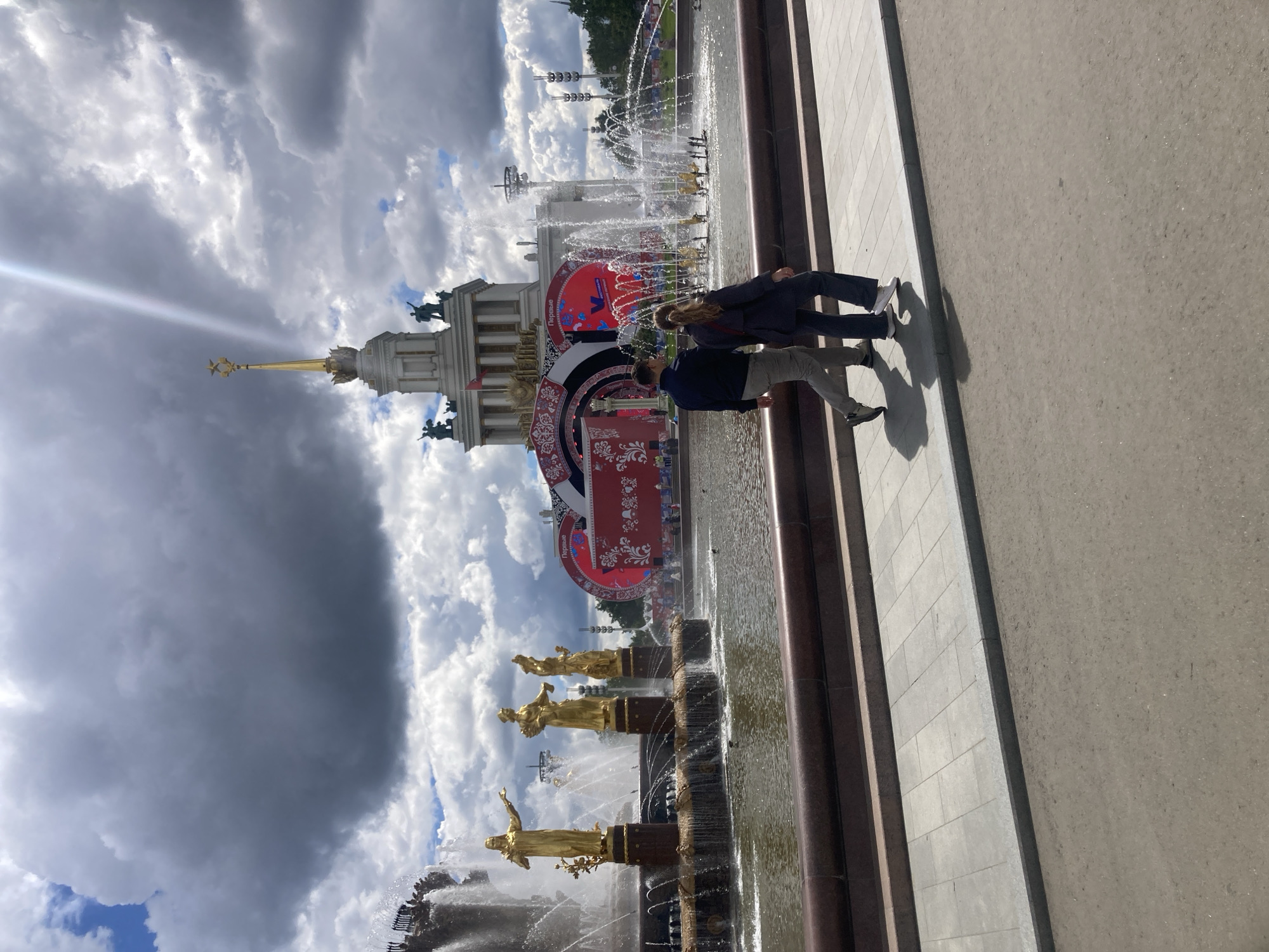 Here's a description of the image:
Two people are standing next to a long, ornate fountain in what appears to be a public space. One person is wearing dark pants and a jacket, while the other is wearing light-colored pants and a dark jacket. Behind them is a large building with a spire topped by a golden star. There are statues of golden women, and large banners are also behind the couple. The sky is cloudy, and the photo is taken from a slightly elevated angle. The image is taken at the Exhibition of Achievements of National Economy (VDNKh) in Moscow, Russia.