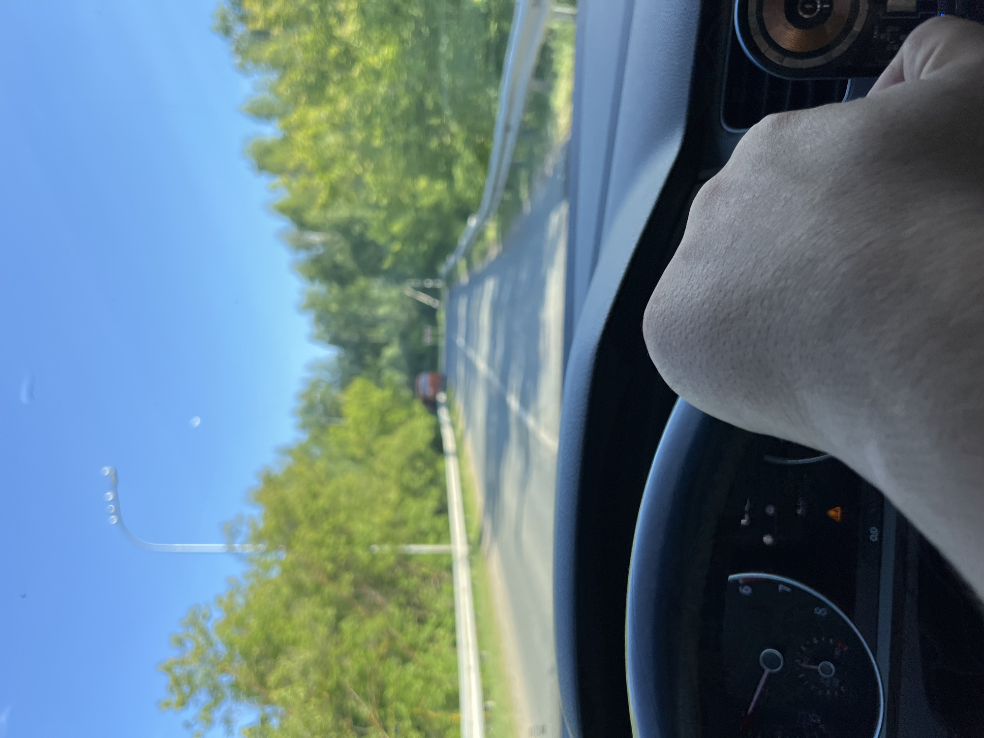 The image shows a point-of-view shot from inside a car driving on a road. A person's hand is visible gripping the steering wheel. The road is paved and lined with guardrails on the right side. Trees and greenery are visible on both sides of the road, with a bright blue sky overhead. In the distance, a red vehicle is seen on the road. The car's dashboard with a speedometer and warning lights is partially visible in the foreground. The scene appears to be in a rural or suburban area. Based on the provided information, this scene is located in the city of Vasilyevo, Russia.