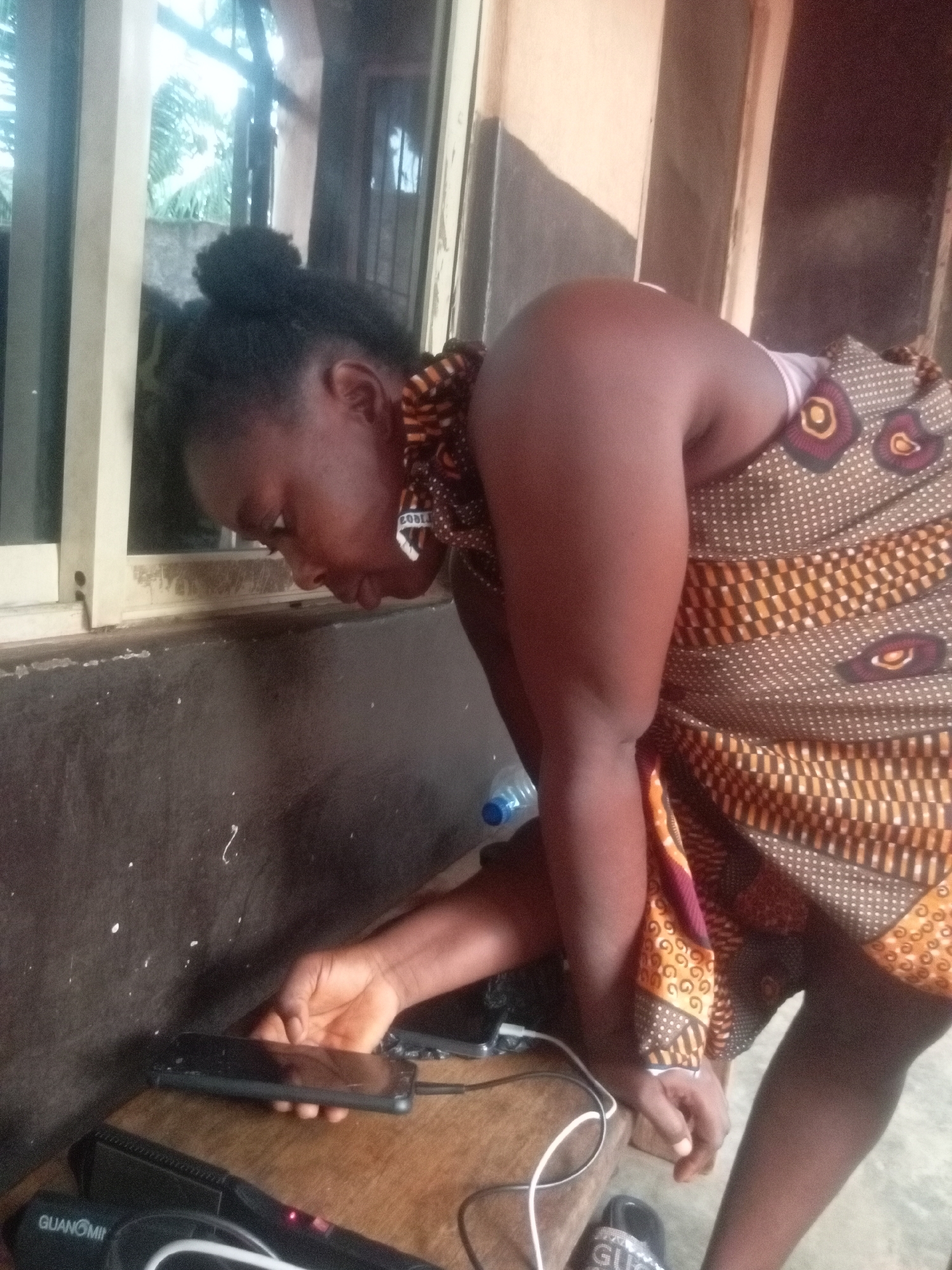 A woman wearing a brown patterned cloth wrapper is leaning over a table indoors, looking at a smartphone in her hand. The phone appears to be charging. A black hair straightening appliance and a water bottle are also on the table. The location is identified as Ogidi, Nigeria.