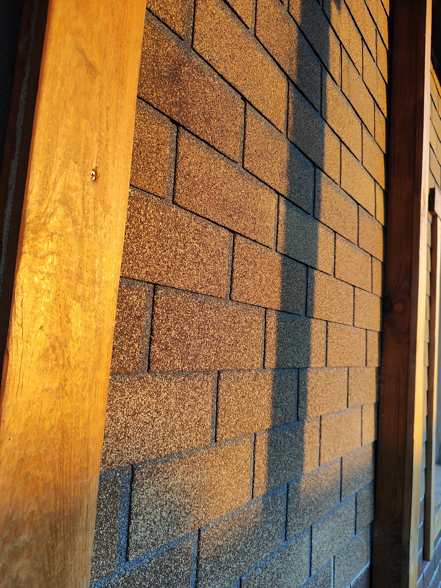 The image shows a close-up, angled view of an exterior wall covered in brown asphalt shingles. A vertical wooden beam, brightly lit by sunlight on its left side, runs along the left edge of the frame. A smaller vertical wooden element is visible on the right edge of the frame. The shingles exhibit a granular texture and are arranged in a staggered pattern. Shadows from an unseen object cast vertical lines across the shingle wall, indicating the direction of sunlight. The scene appears to be an outdoor construction or renovation site. The location is identified as Marusino, Russia.